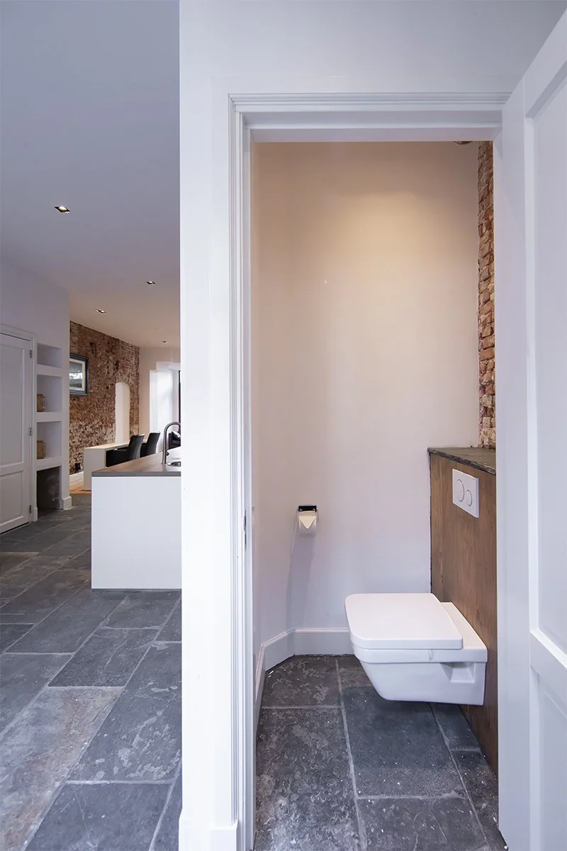 Modern toilet in a small, partially enclosed bathroom with grey tile flooring, white walls, and exposed brick detail.