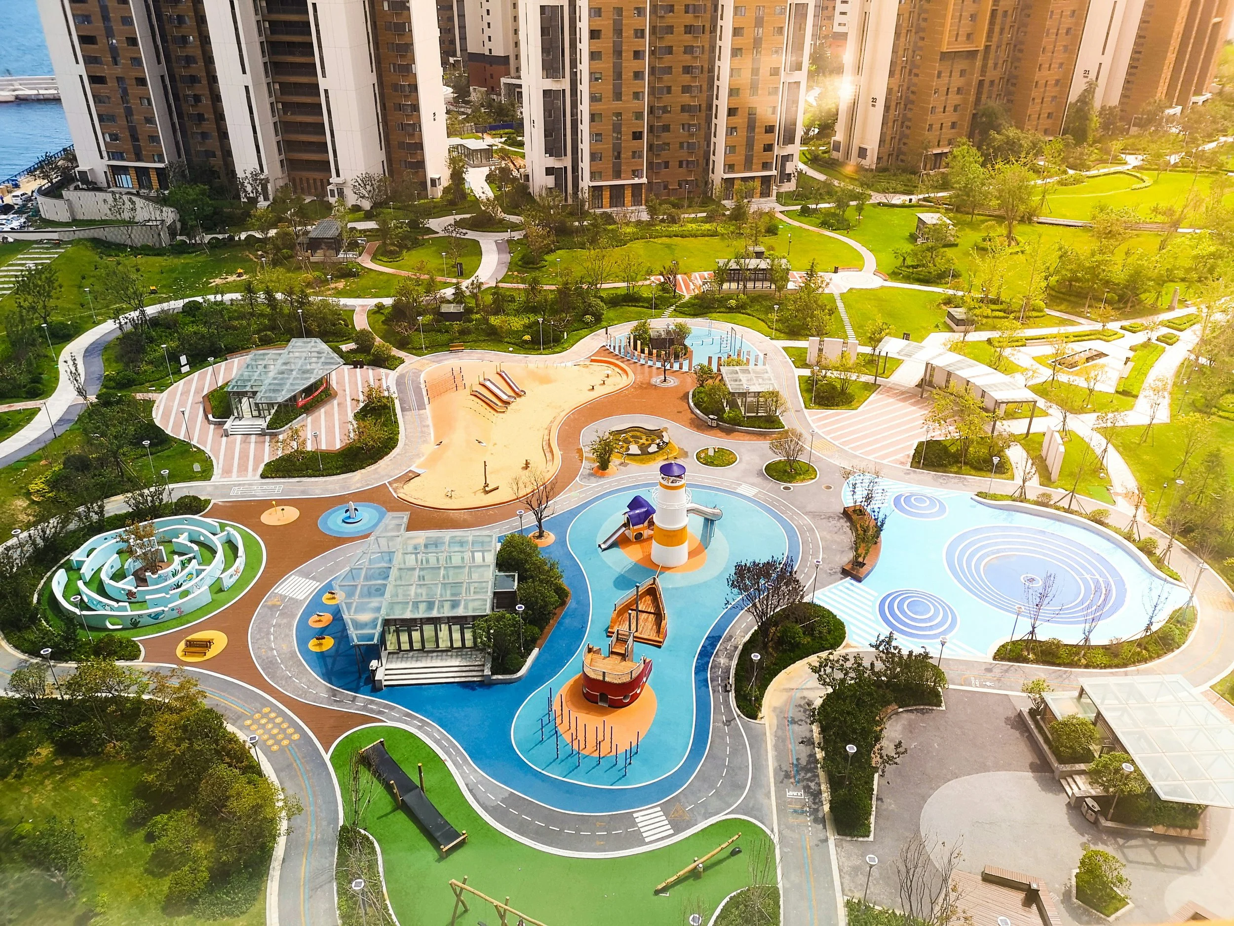 Aerial view of a colorful children's playground and park in an urban area with high-rise residential buildings, walking paths, green lawns, trees, and landscaped gardens.