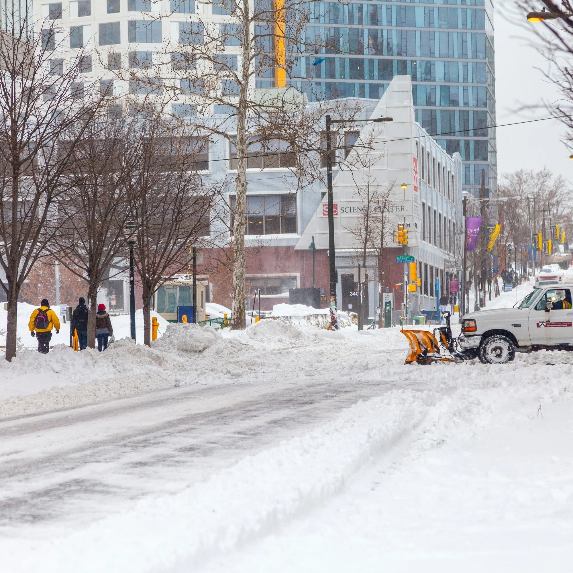 Snow-covered city street with people walking, snowplow, and tall modern buildings in the background.