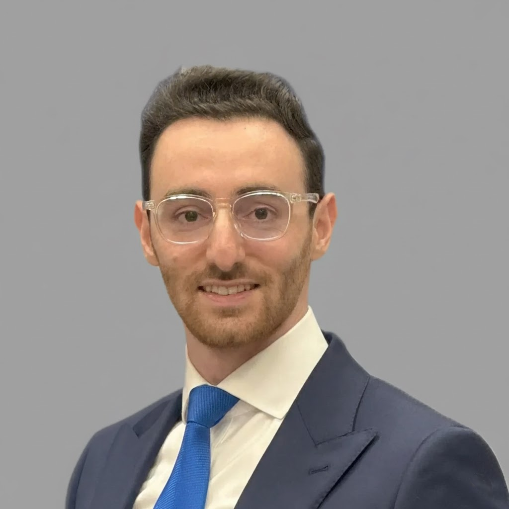 A man with glasses in a business suit smiling against a plain gray background.