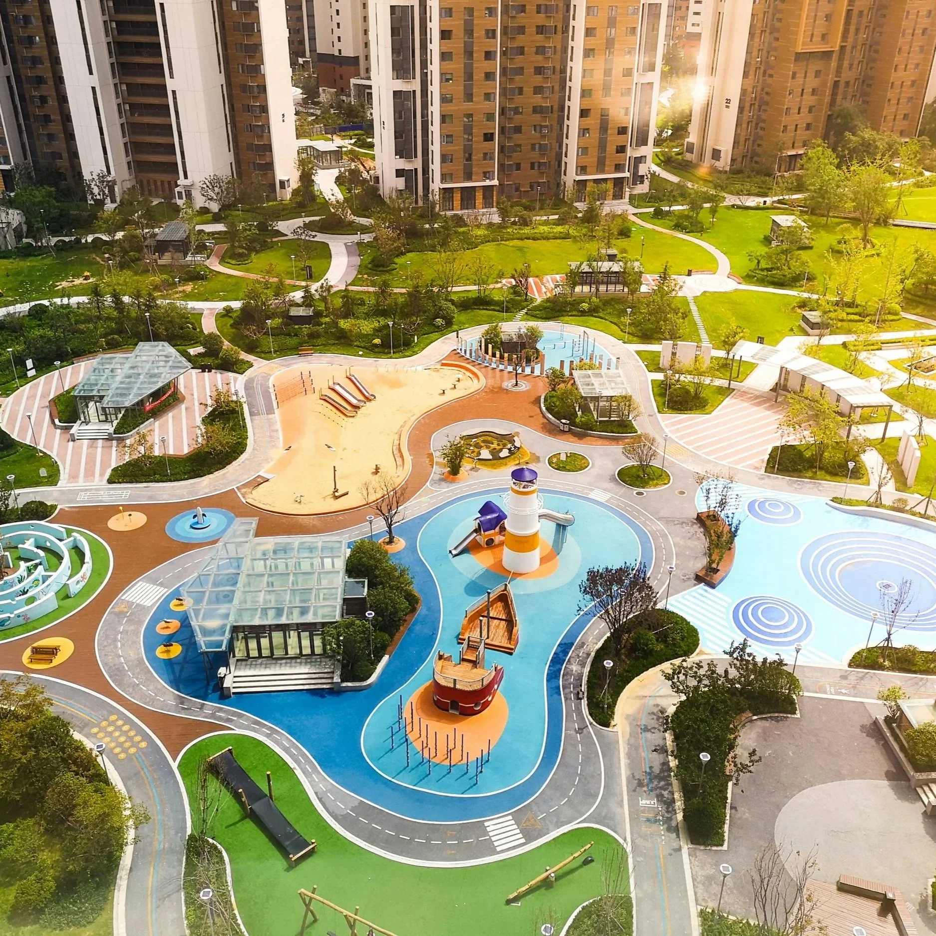 Colorful children's playground with various play structures, slides, a lighthouse, and open spaces, surrounded by high-rise residential buildings and green lawns.