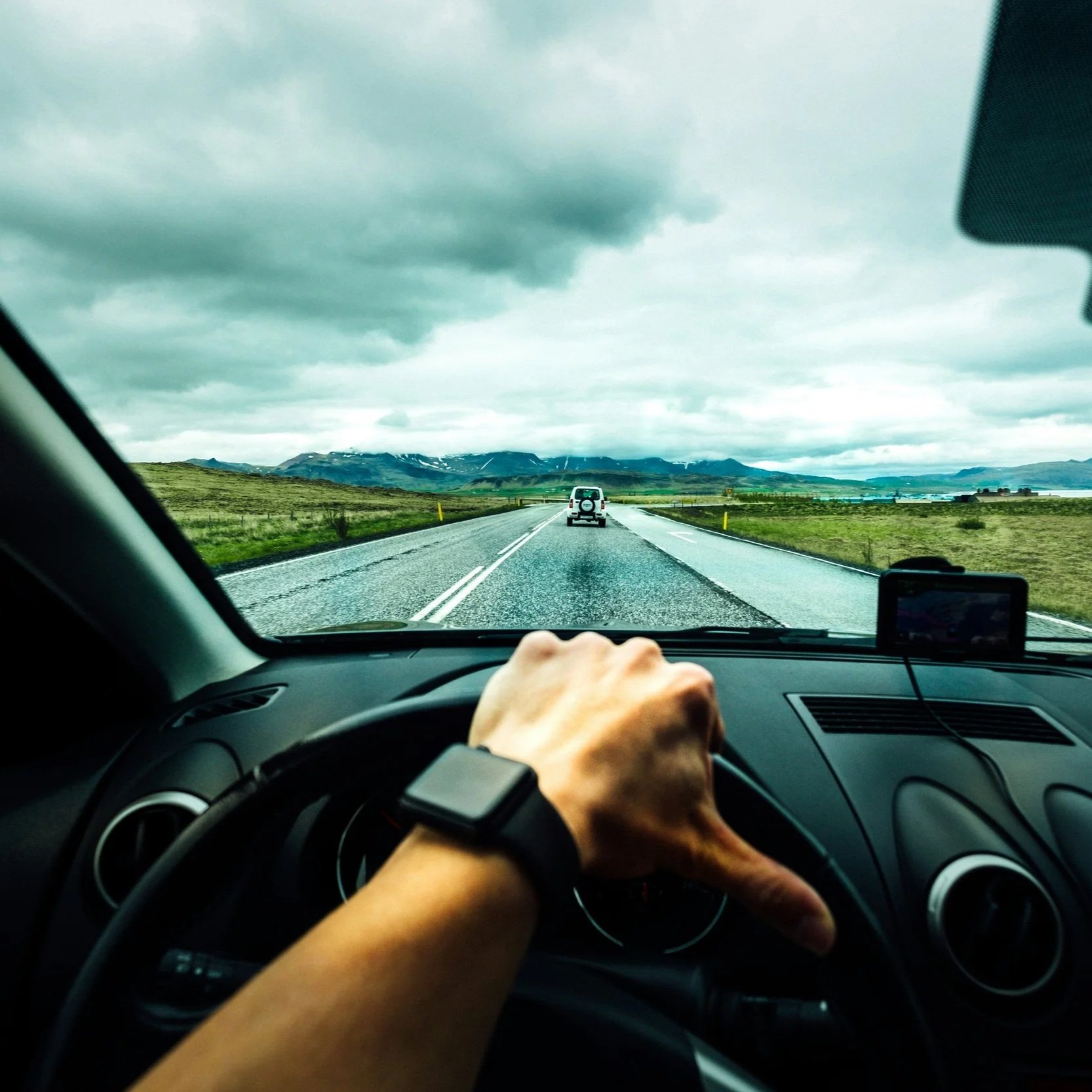 View from inside a car showing a driver’s hand on the steering wheel, traveling on a two-lane highway through a green landscape with mountains in the background under a cloudy sky.