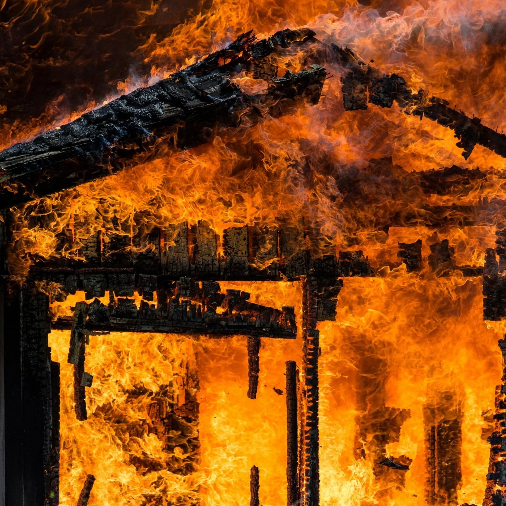 A house engulfed in bright orange and yellow flames, with charred remains of the structure visible through the blaze.