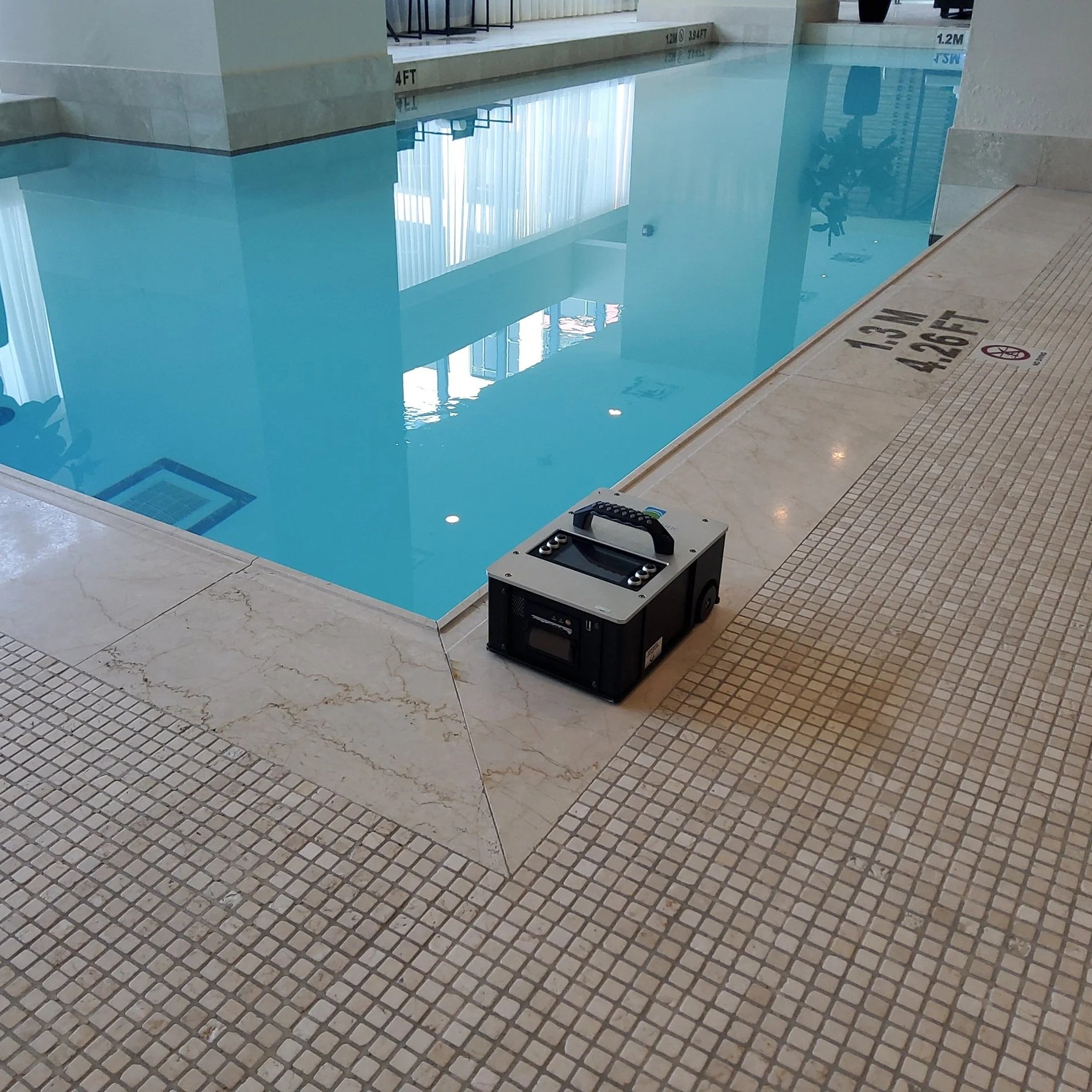 A black and gray portable electronic device, possibly a radio or speaker, resting on a tiled floor next to an indoor swimming pool.