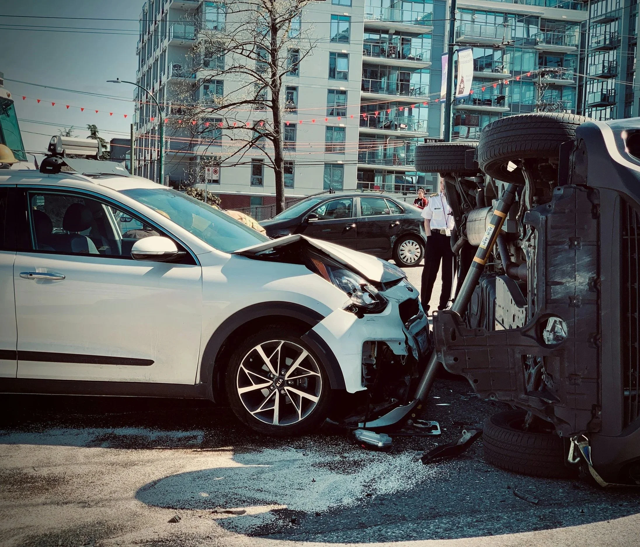 A car accident involving a white car and an overturned black vehicle on a city street with high-rise buildings in the background.