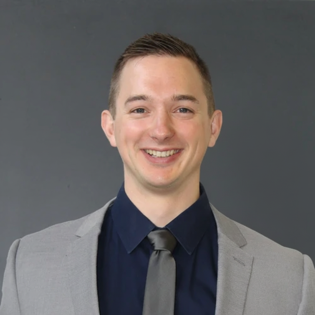 A smiling young man in a gray blazer, dark shirt, and gray tie posing against a dark background.
