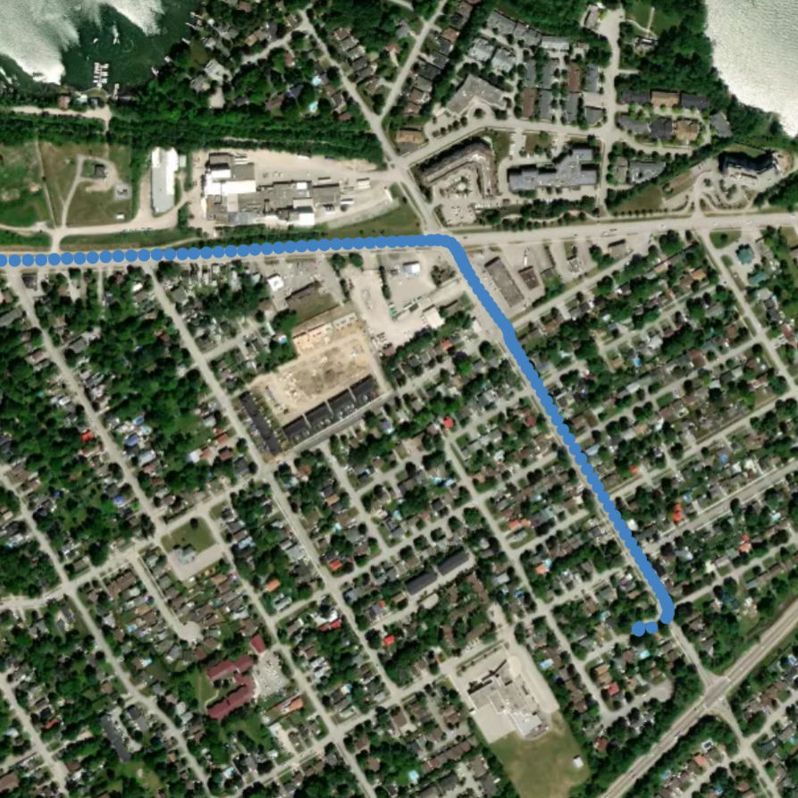 An aerial view of a residential neighborhood with houses, streets, green trees, and a large building. A blue line traces a route through the neighborhood.