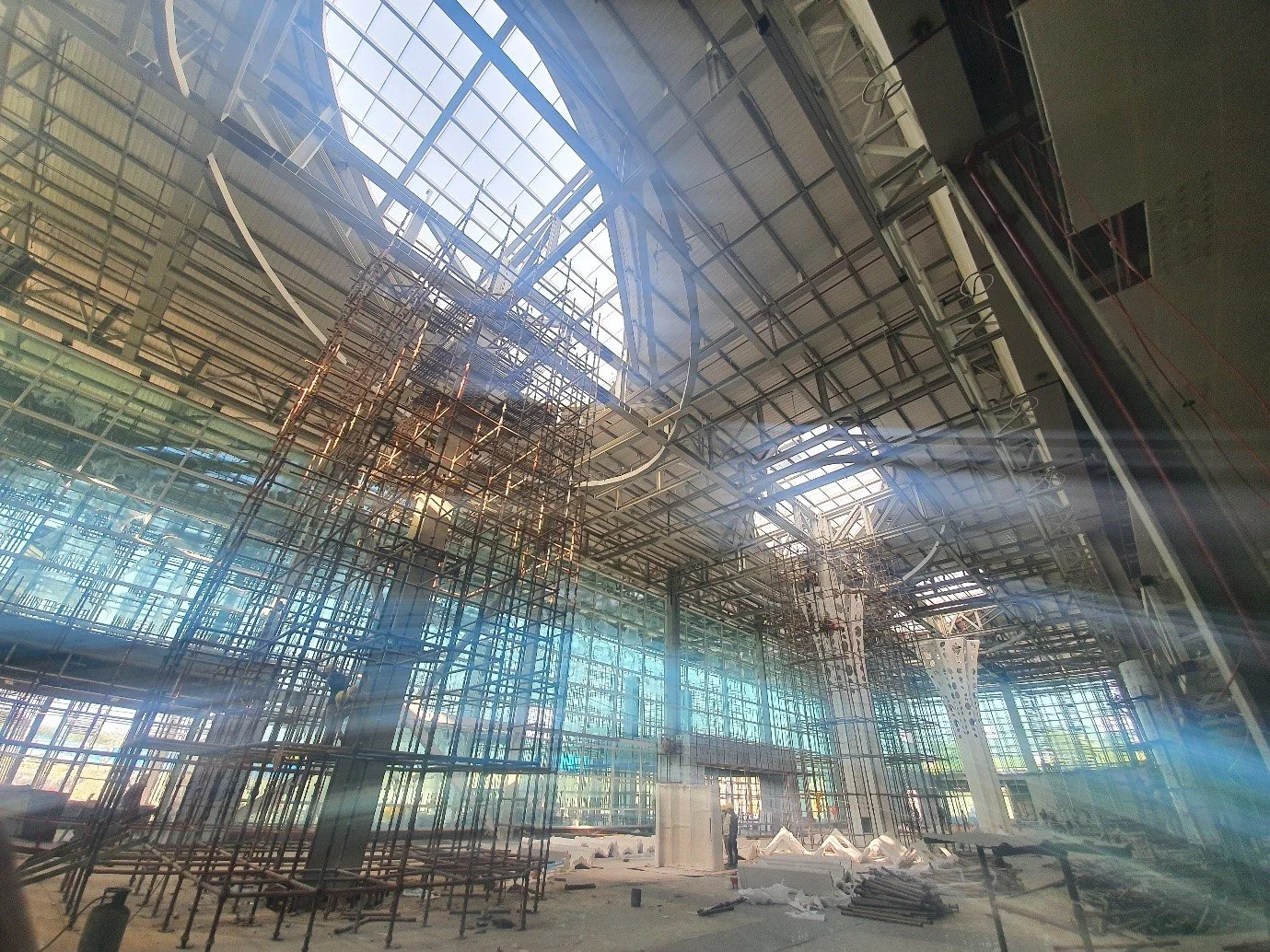 Under construction interior with scaffolding and steel framework in a large building, sunlight streaming through glass ceiling panels.