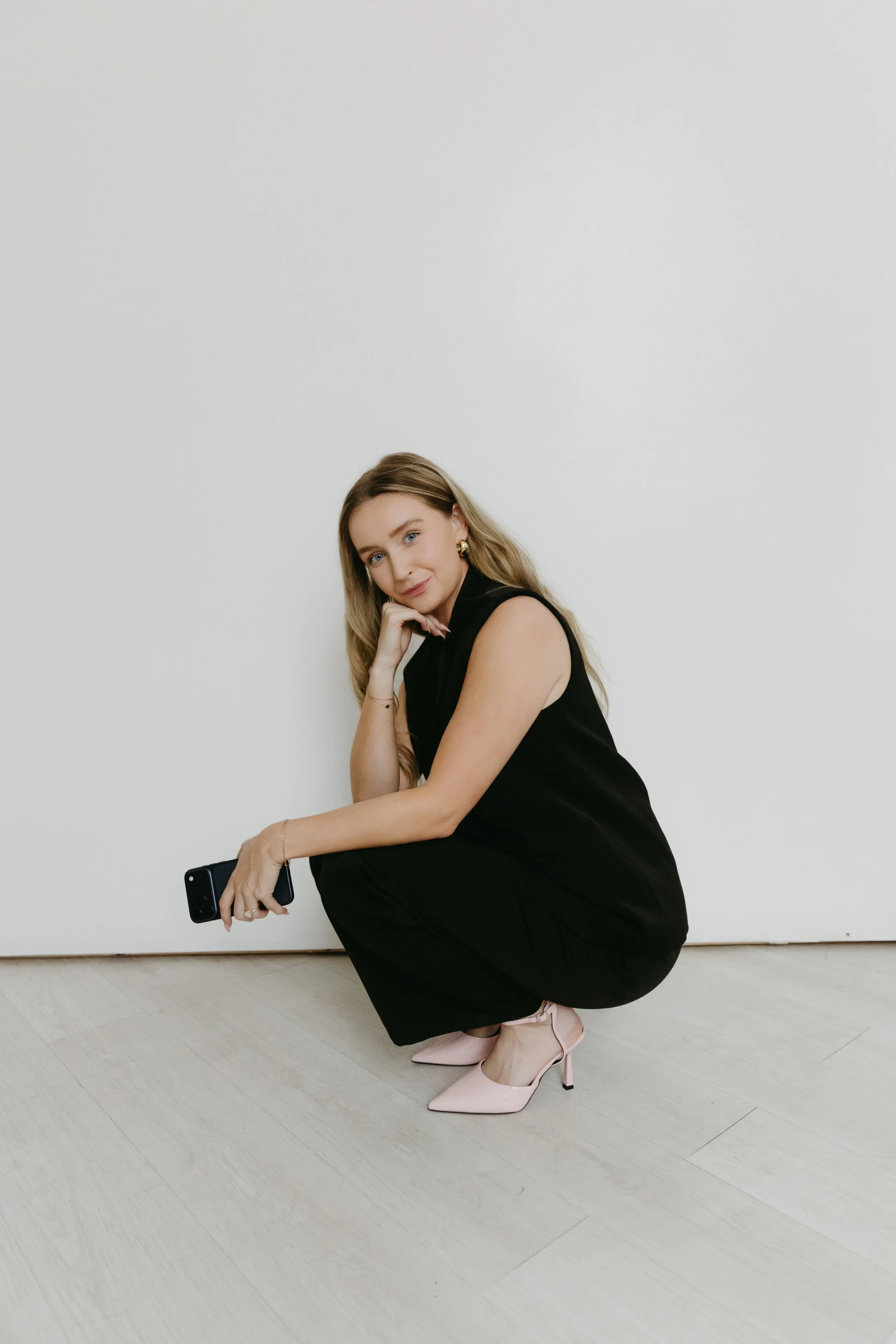 A woman wearing a sleeveless black top and black pants squatting on a light-colored wooden floor, holding a smartphone in her right hand, with long blonde hair, pink high heels, and a neutral-colored background.