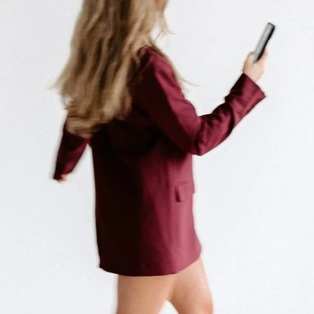 A woman with long hair wearing a burgundy blazer and matching skirt holding a smartphone against a plain white background.