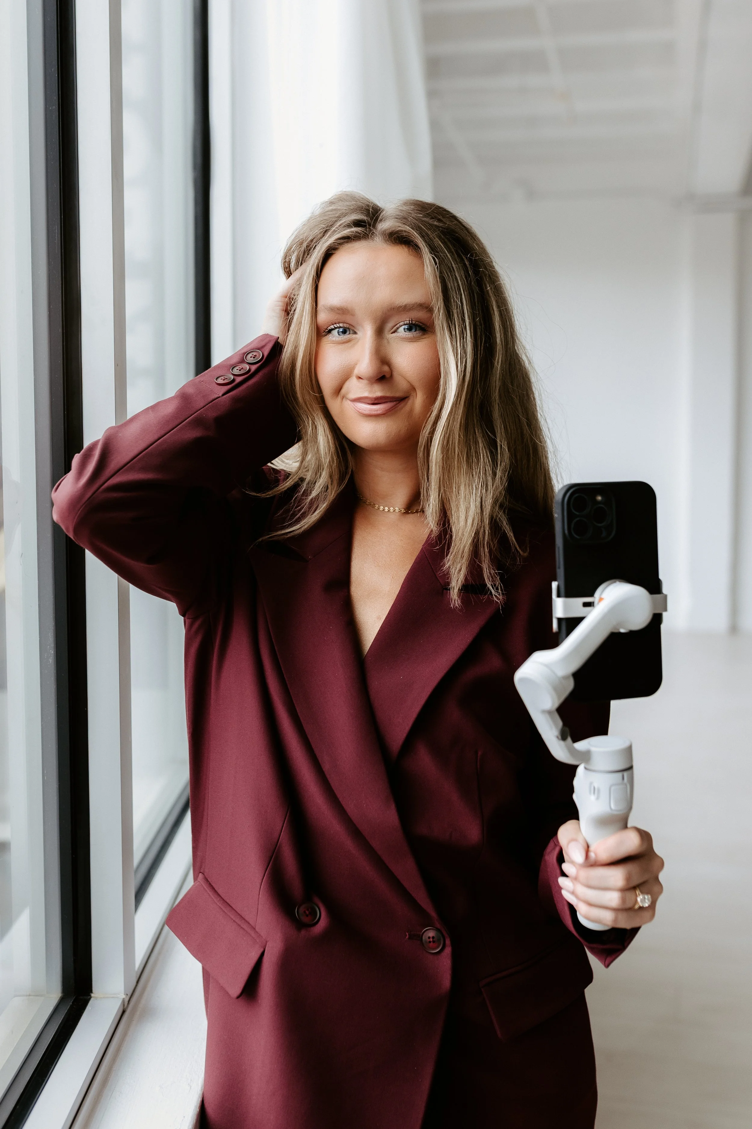 A woman with blonde hair and a burgundy blazer takes a selfie using a smartphone mounted on a gimbal, standing near large windows in a bright room.
