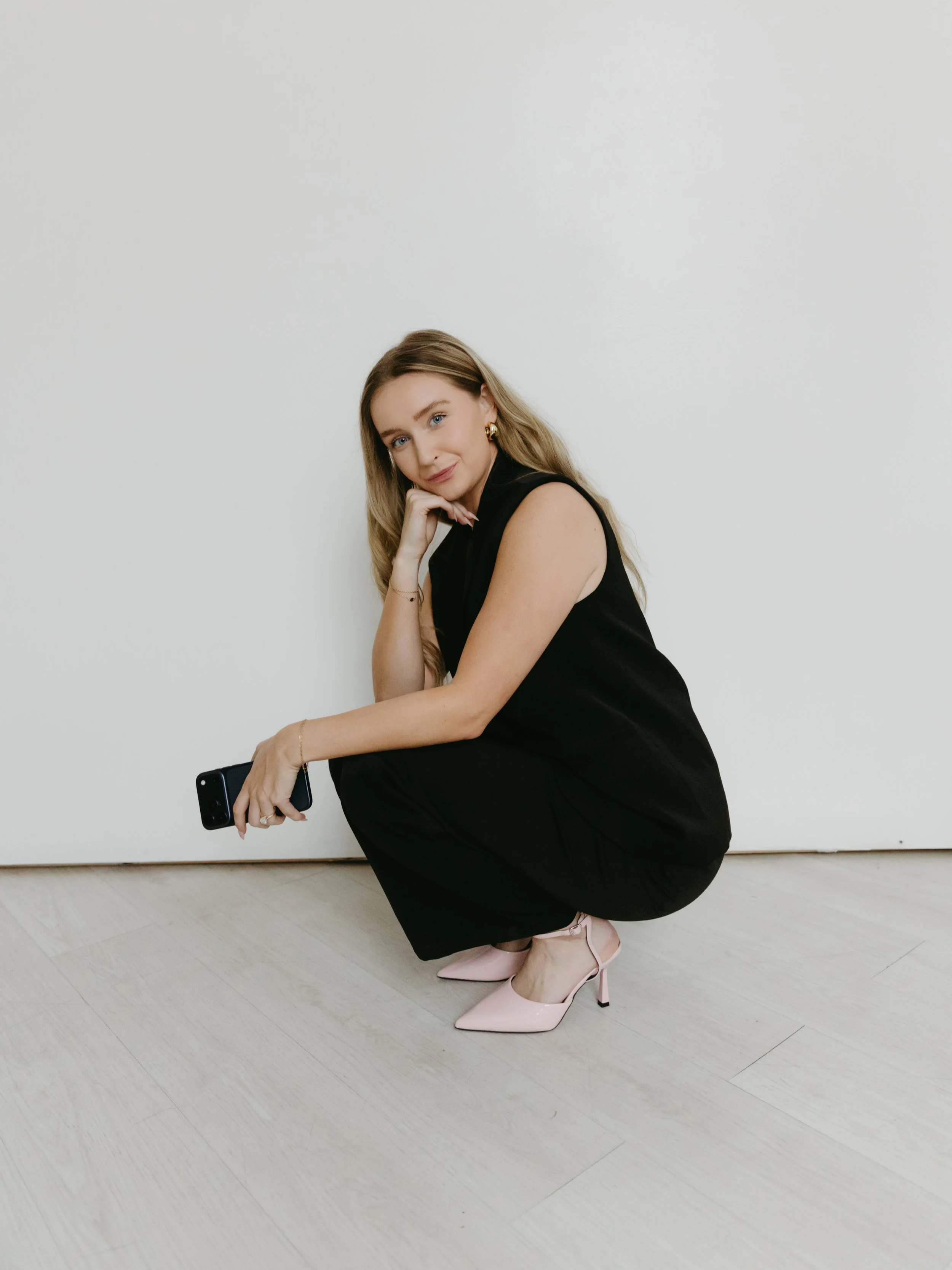 A woman with long blonde hair squatting on the floor next to a plain white wall, wearing a black sleeveless jumpsuit and pink high heels, holding a smartphone in her right hand and resting her chin on her left hand.