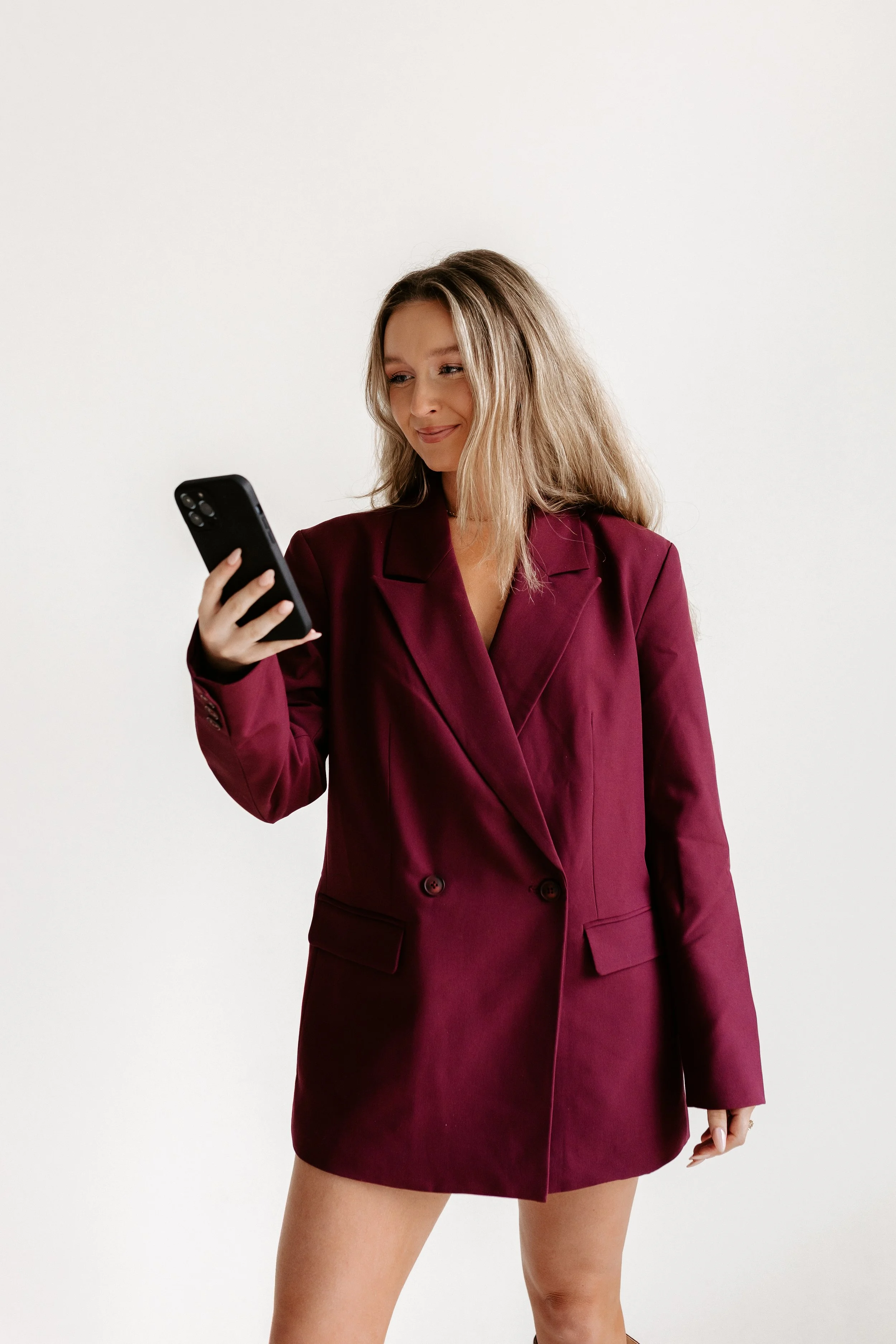 A woman with blonde hair wearing a burgundy blazer dress is looking at her phone and smiling.