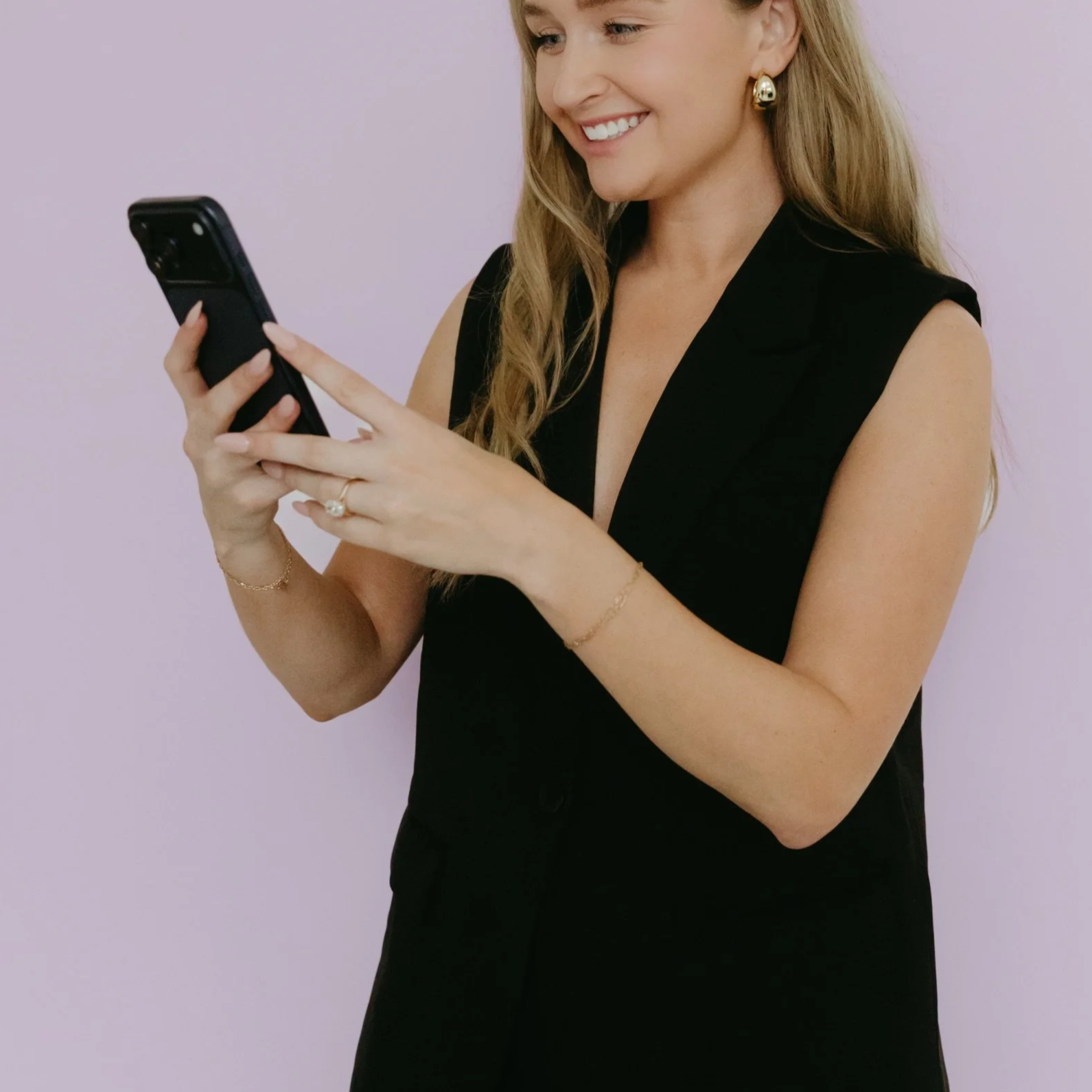A smiling woman with long blonde hair wearing a black sleeveless dress, gold earrings, and minimal jewelry, looking at her phone against a light purple background.