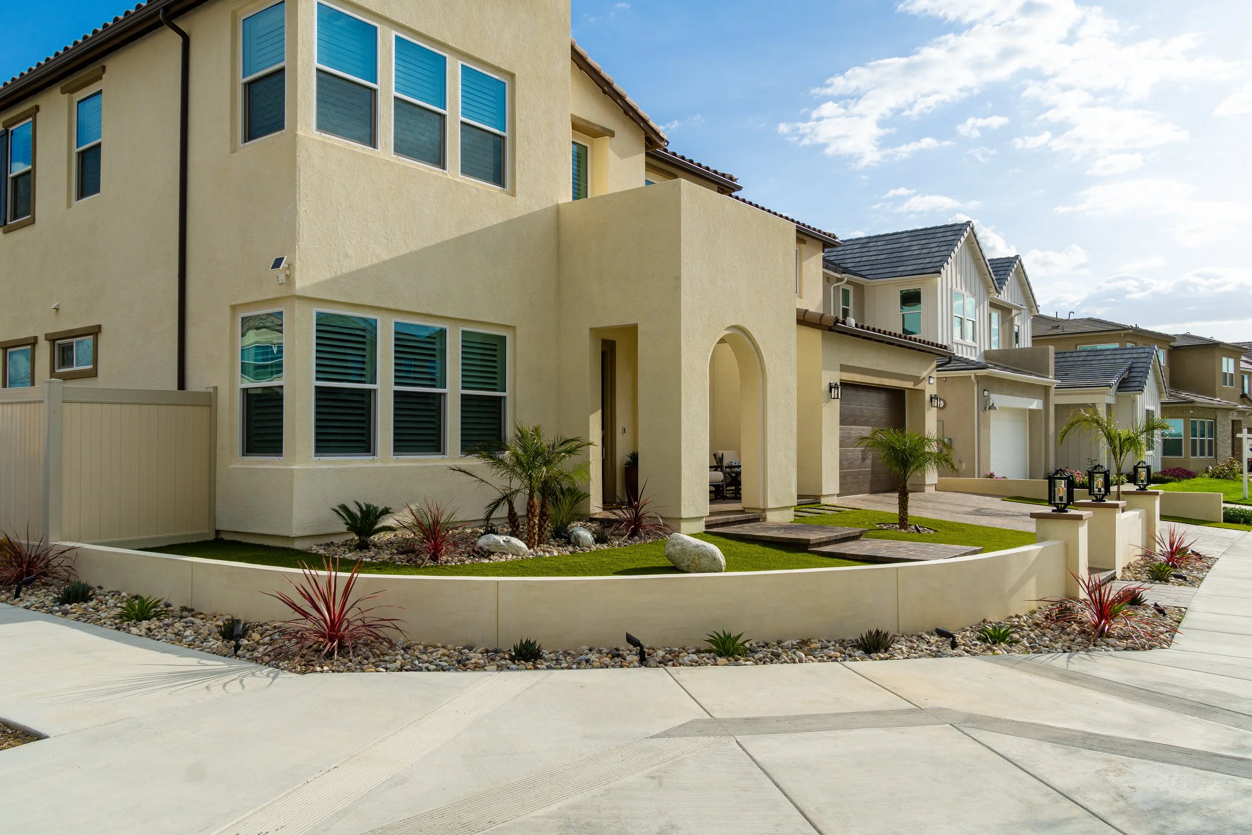 Modern two-story house with a beige stucco exterior, multiple square windows with shutters, and a landscaped front yard with palm trees, rocks, and plants. There is a curved sidewalk and a driveway leading to the garage.