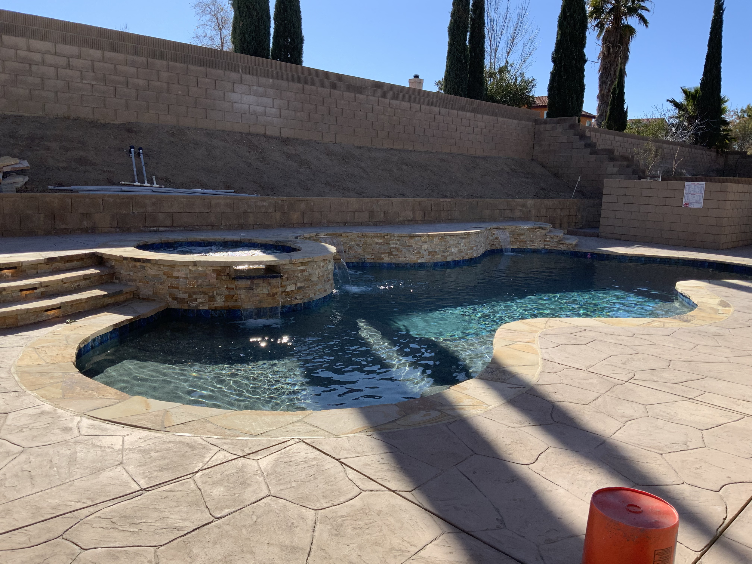 A backyard swimming pool with a curved design, a spa with a stone exterior, and a water feature flowing into the pool, surrounded by a stamped concrete deck and a retaining wall with steps, with trees and a blue sky in the background.