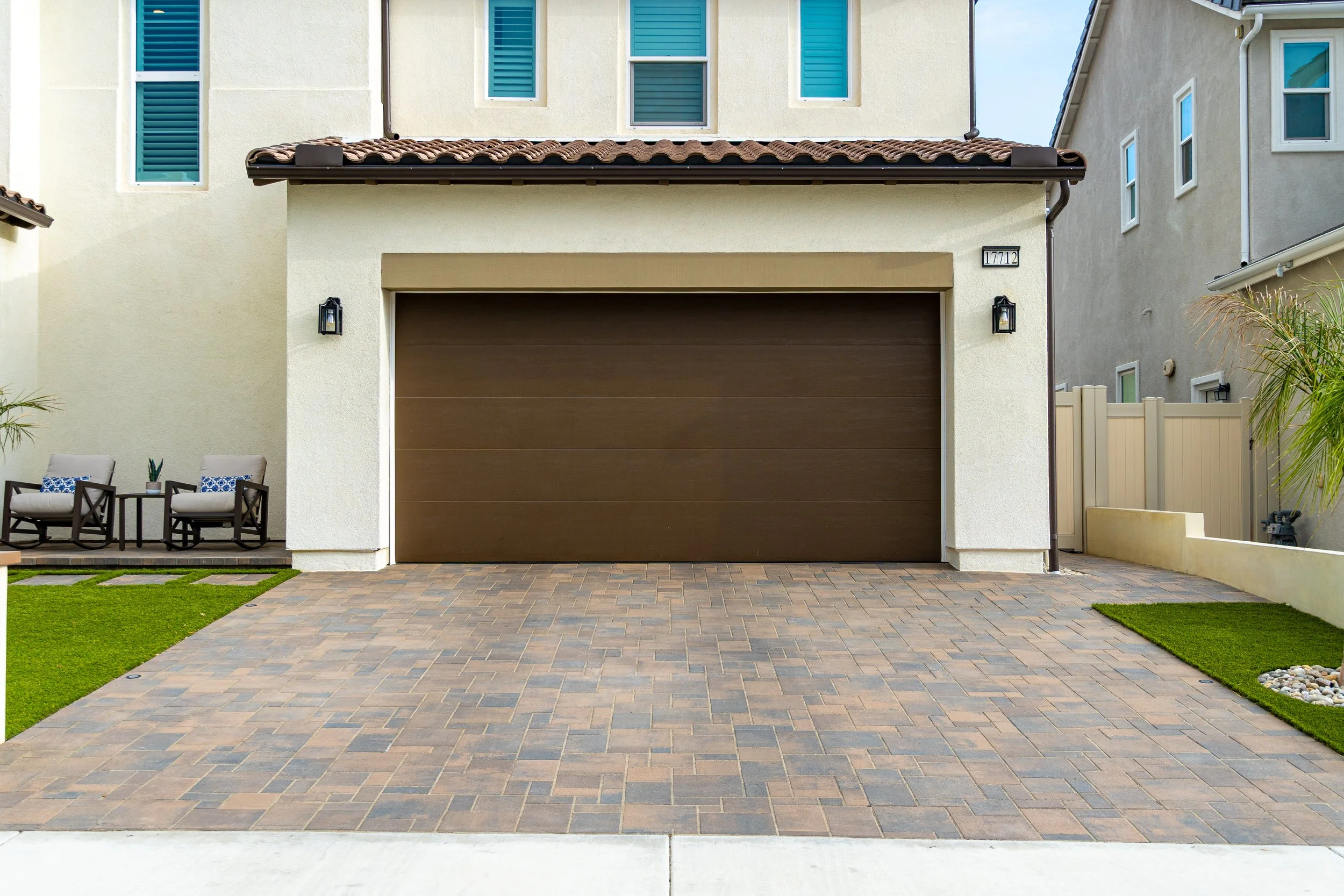 Front view of a modern house with a brown garage door, beige exterior walls, two seating chairs with small tables on the left, a small palm tree, and a pathway with pavers leading to the garage.