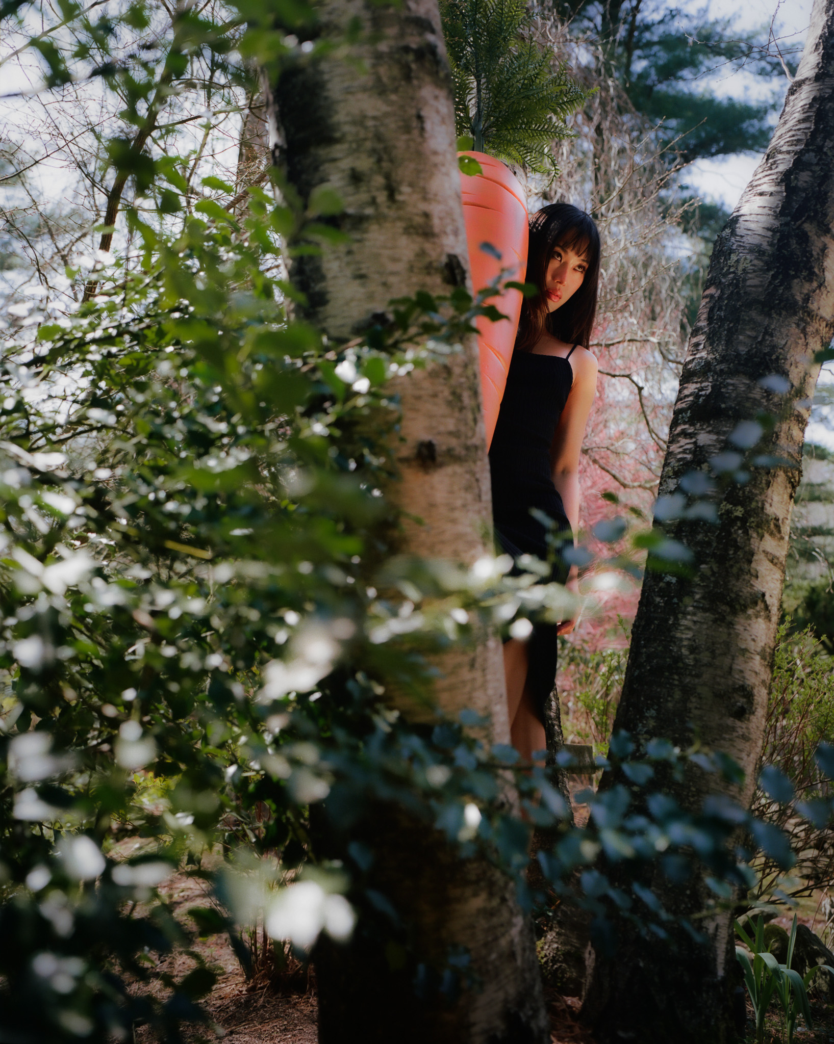 A young woman with dark hair and a black top leaning against a tree in a forested area with sunlight filtering through the trees.