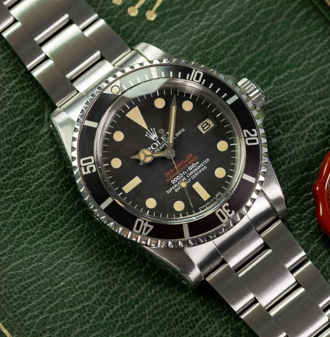 A silver Rolex Sea-Dweller Submariner watch with a black dial, silver bezel, and metal band, lying on a textured green surface.