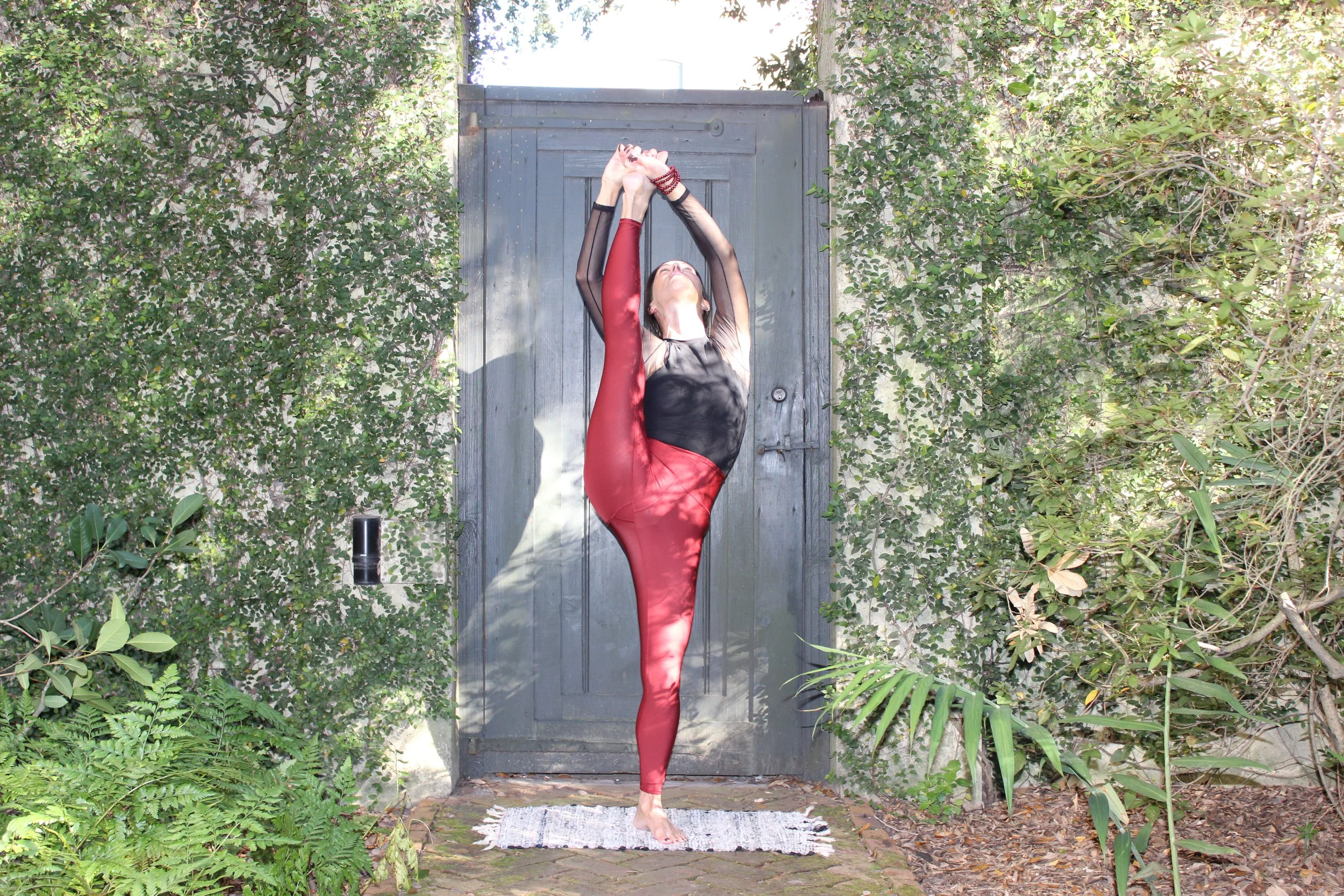 A woman practicing yoga outdoors, performing a standing split pose with one leg raised high and hands holding the foot, in front of a gray wooden gate surrounded by lush green foliage.