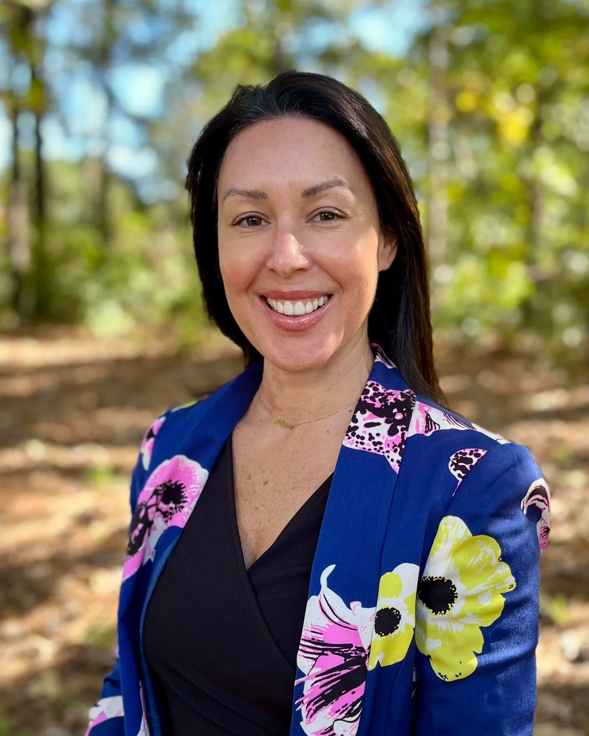 A woman with medium-length black hair, smiling, wearing a black top and a blue blazer with pink, yellow, and black floral patterns, standing outdoors in a forest setting with trees and sunlight filtering through leaves.