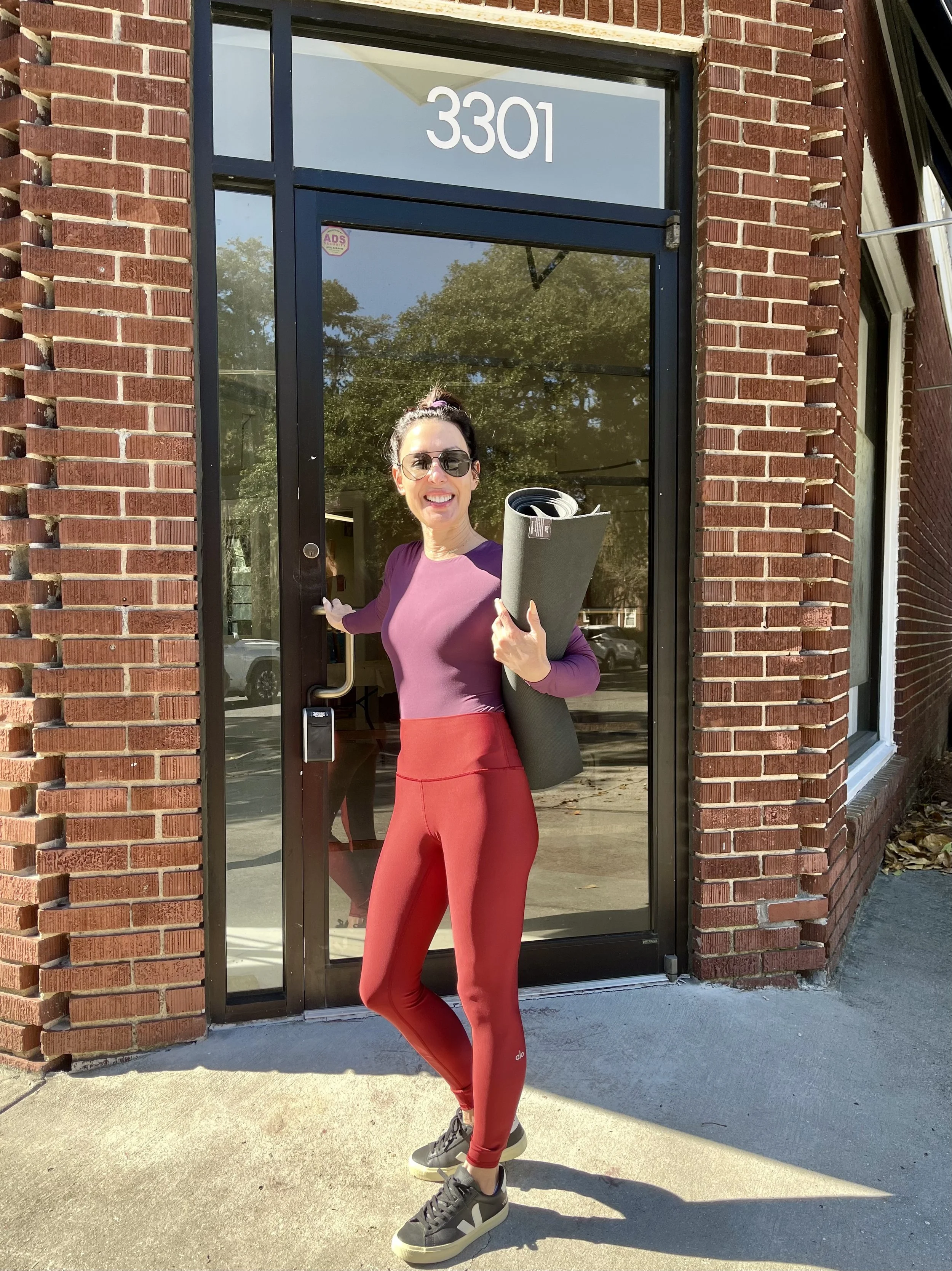 A woman standing outside a glass door with the number 3301, holding a yoga mat rolled up under her arm, dressed in athletic wear, smiling, and wearing sunglasses.