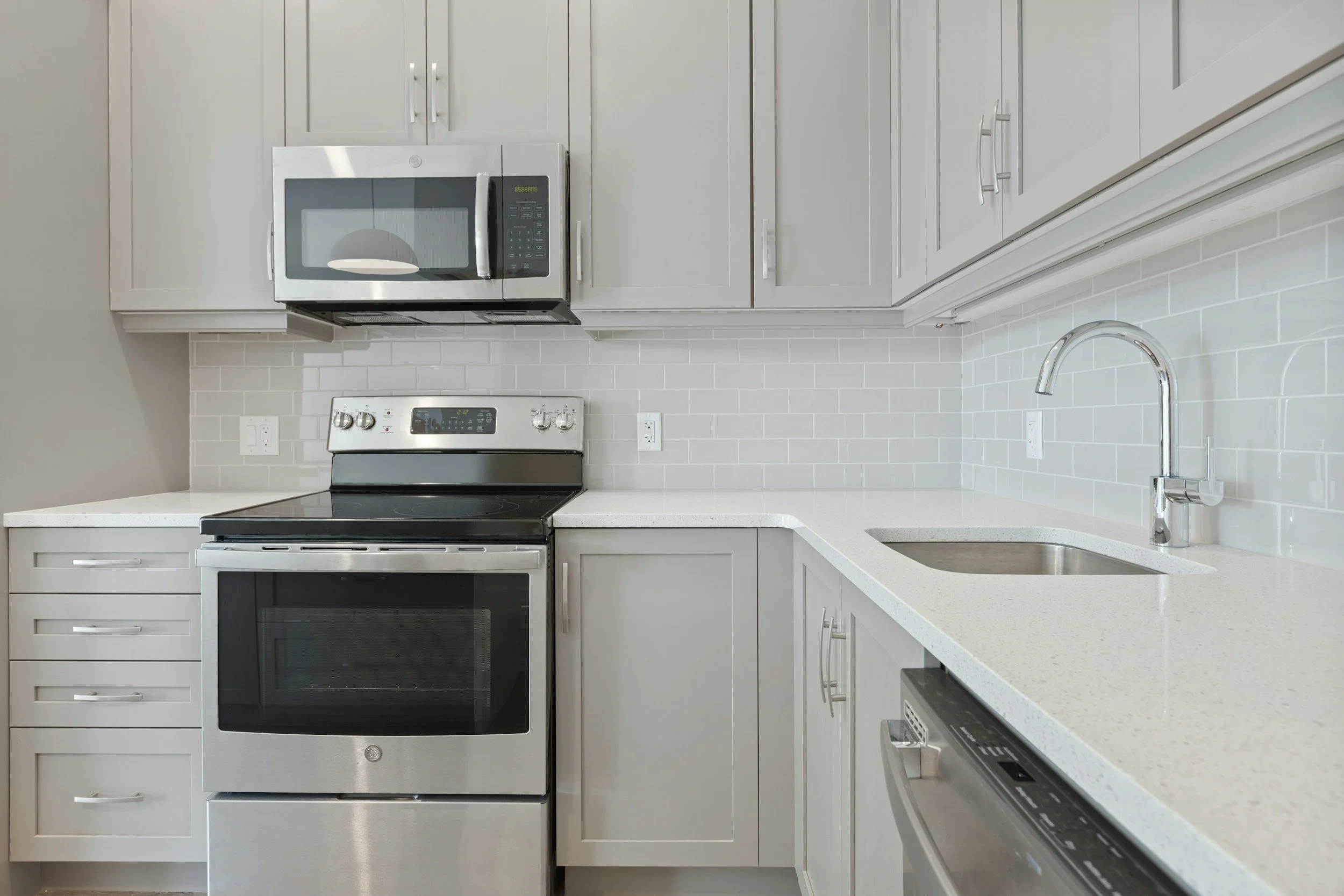 Modern kitchen with white cabinets, stainless steel microwave, oven, and dishwasher, along with a white countertop and light gray tile backsplash.