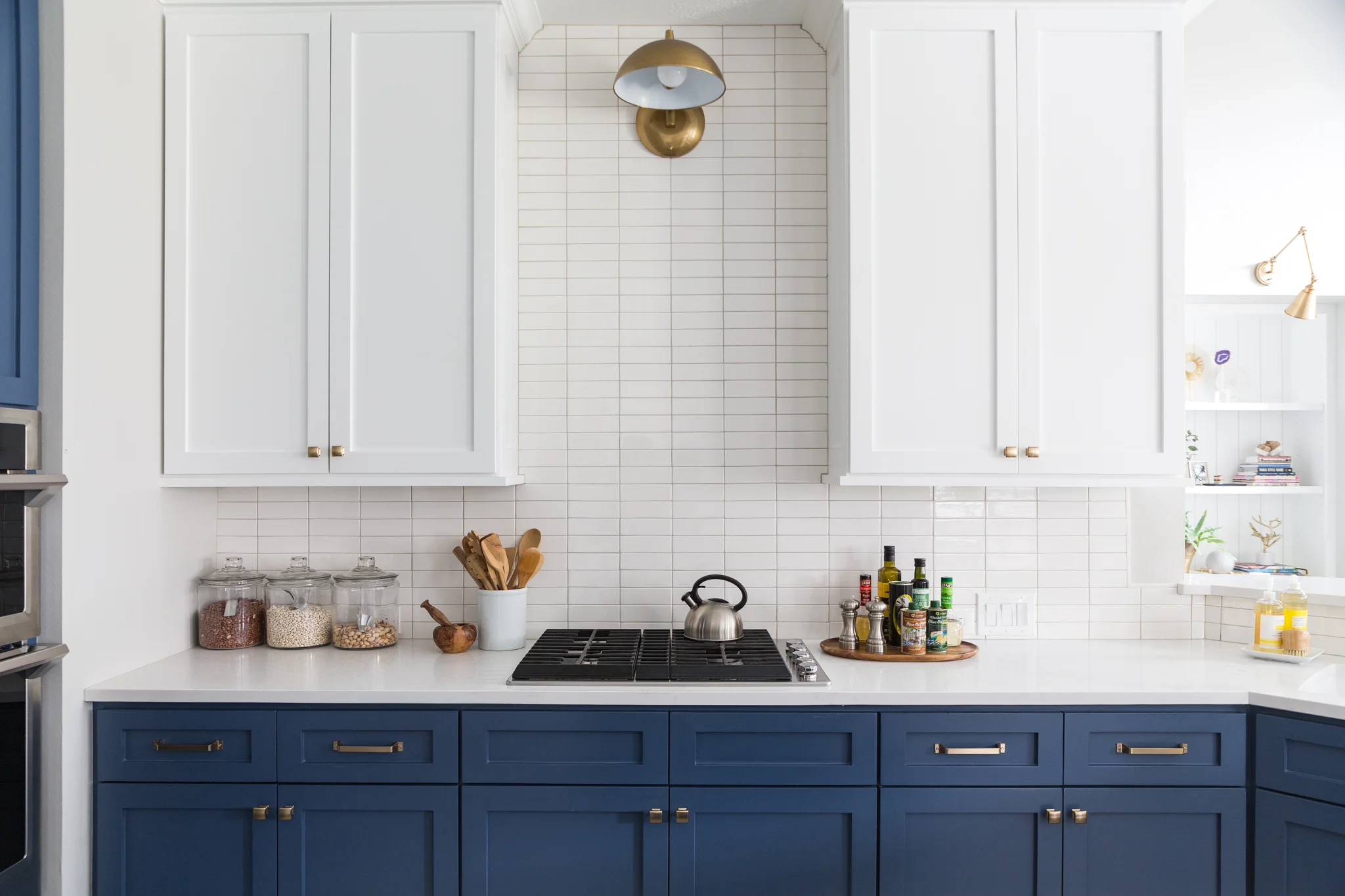 Modern kitchen with white upper cabinets, blue lower cabinets, a white countertop, and a white tile backsplash. A silver kettle sits on a stovetop, and a tray of spices and oils is on the right. Decorative jars and kitchen utensils are on the left, with a gold and white wall lamp above.