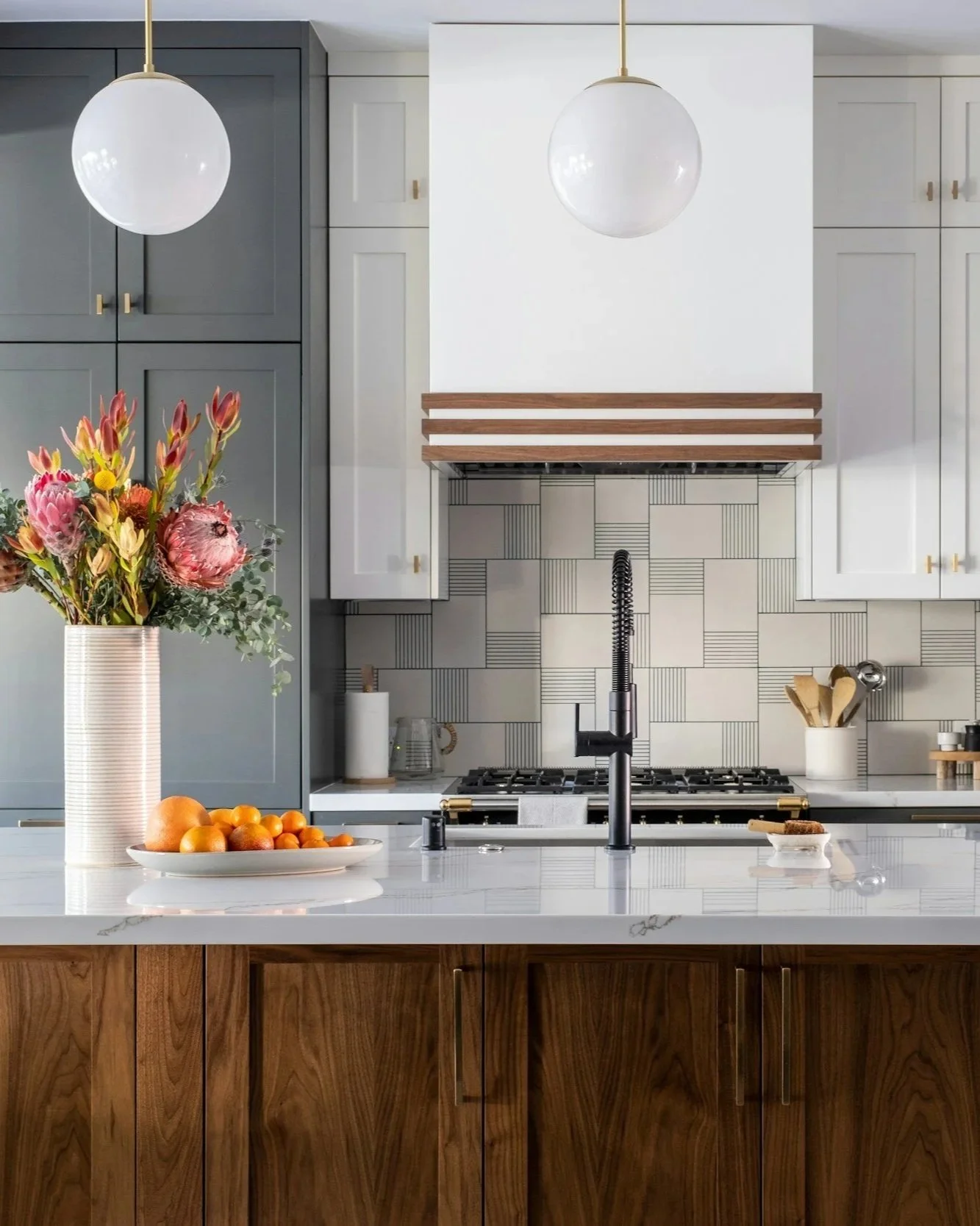 Modern kitchen with grey and white cabinets, a black faucet, a white marble countertop, floral arrangement, and a bowl of fruit.