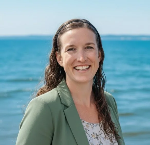 A woman with long brown hair, wearing a green blazer and patterned blouse, smiling at the camera with the ocean and blue sky in the background.