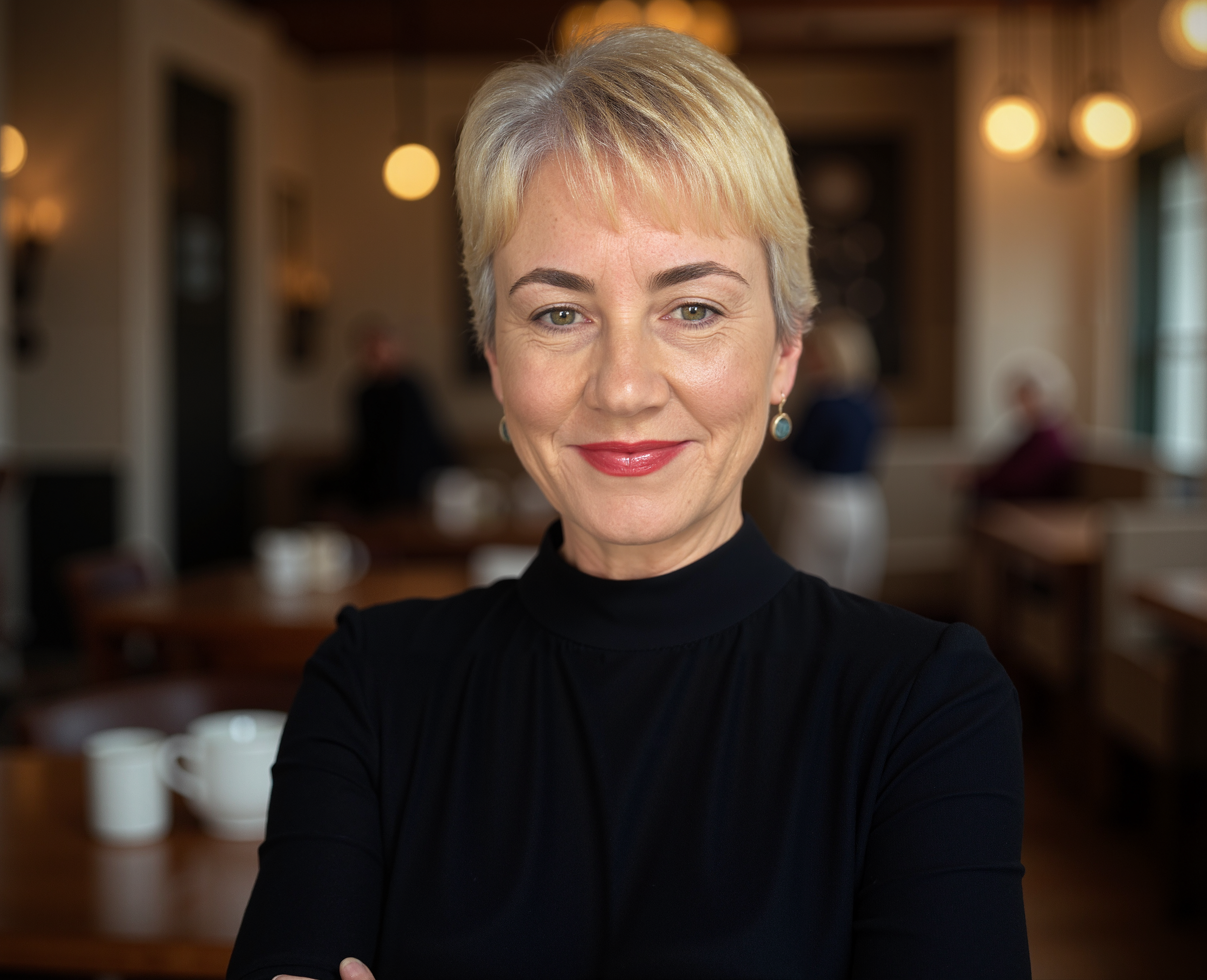 A woman with short blonde hair and wearing a black top, smiling. She is in a cozy, warmly lit room.