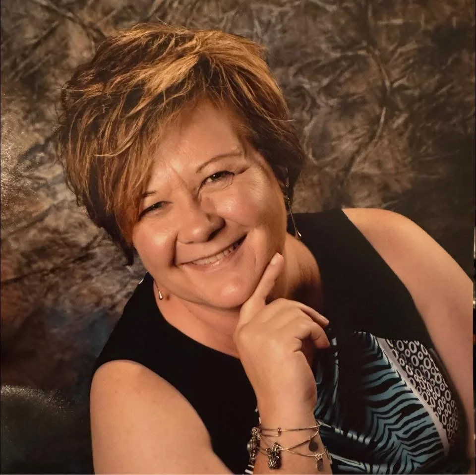 A woman with short, layered brown hair smiling while resting her chin on her hand, wearing a black sleeveless top with a patterned scarf, standing in front of a textured stone wall.