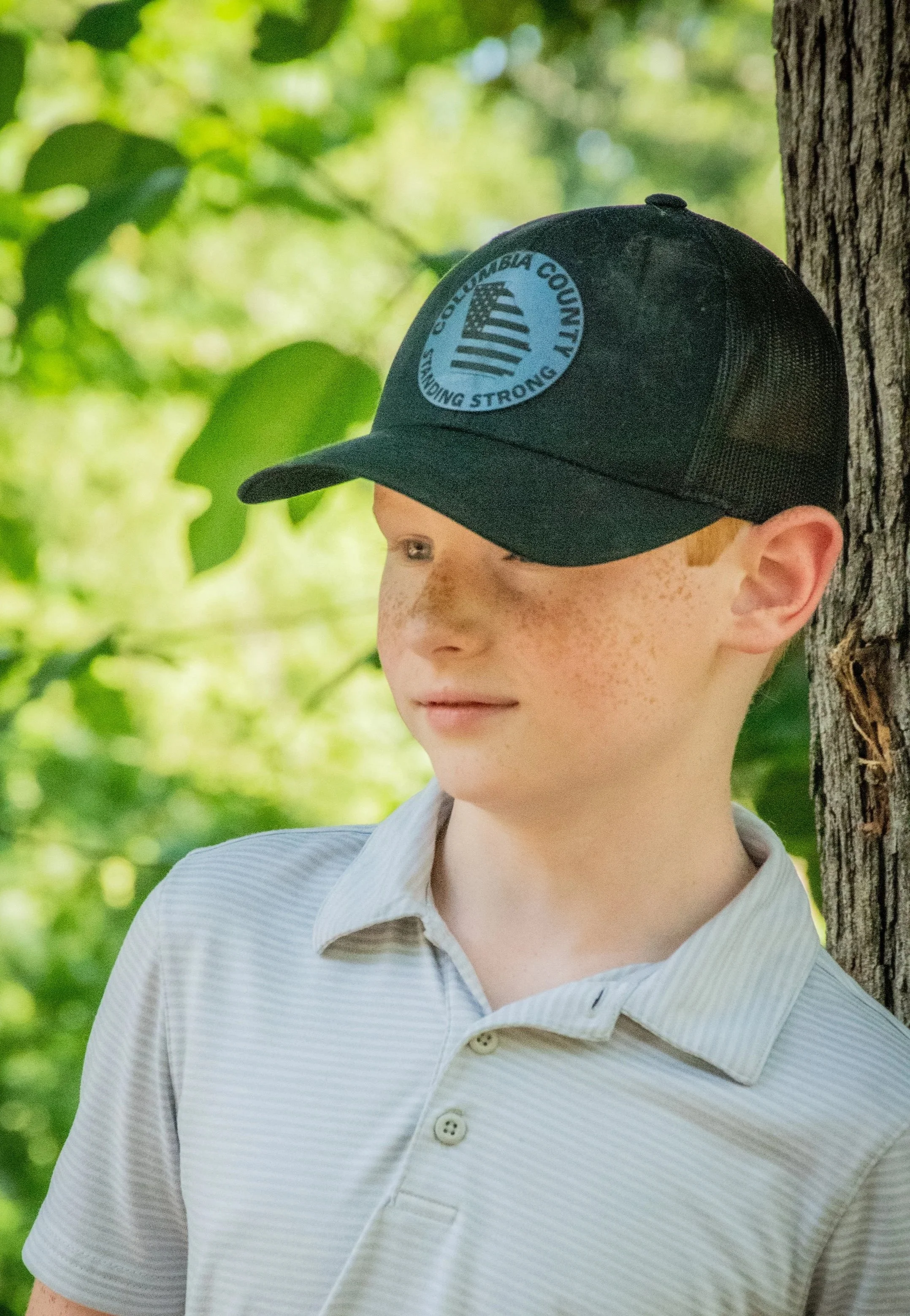 A young boy with freckles and red hair wearing a black Columbia County cap and a light-colored striped collared shirt, standing outdoors next to a tree with green foliage in the background.