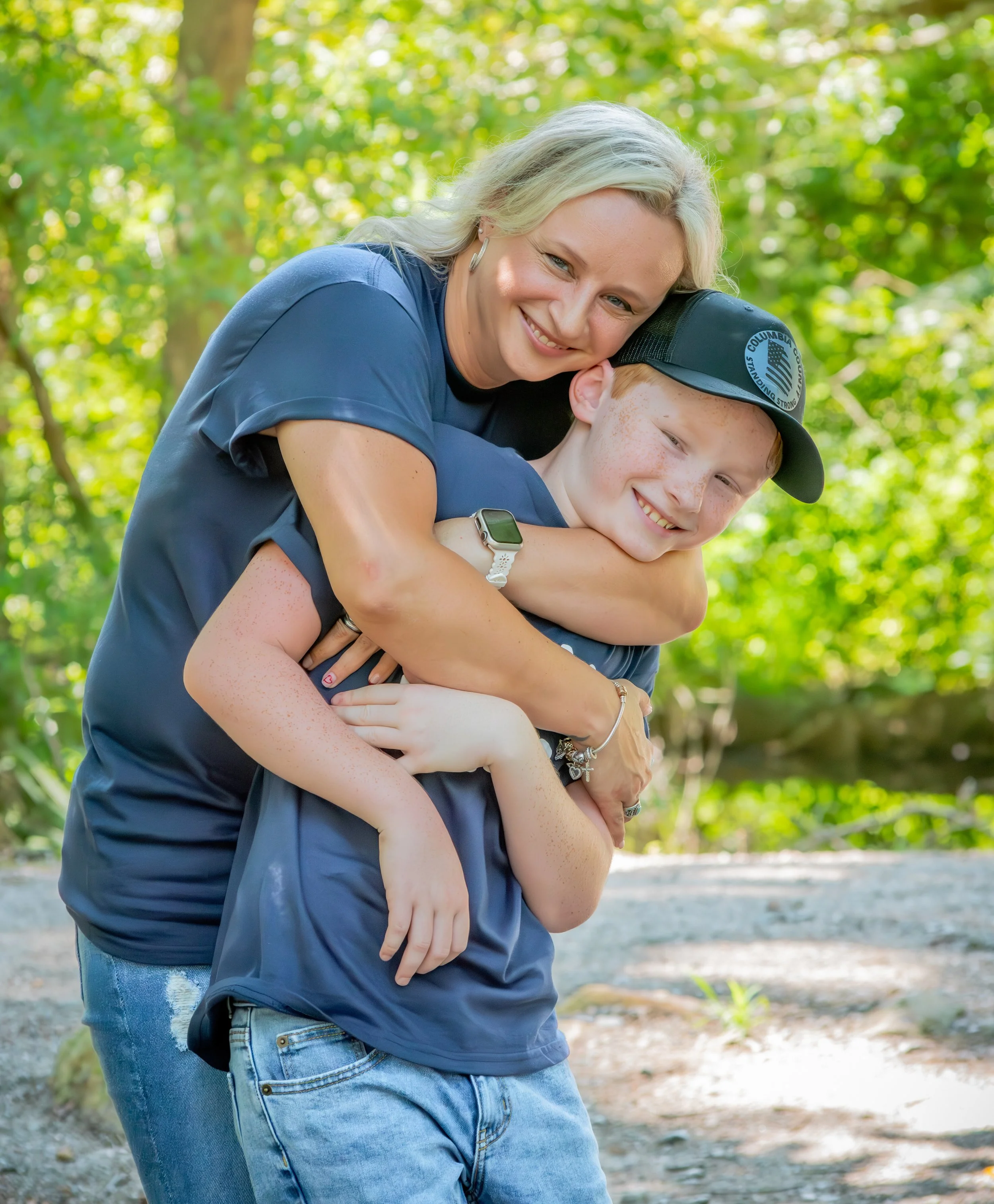 A woman hugging a young boy outdoors in a lush green forest, both smiling.