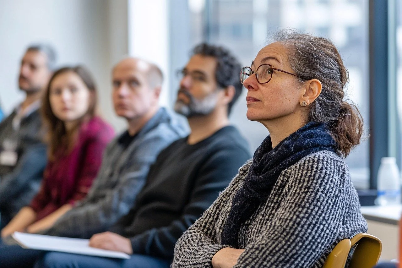 A group of five people sitting in a conference room, listening attentively during a meeting. The focus is on an older woman with glasses and gray hair, sitting with her arms crossed, wearing a patterned blazer and a scarf. The other four individuals are blurry in the background.