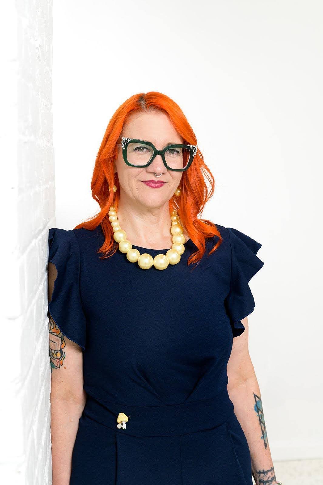 A woman with bright orange hair, green glasses with rhinestones, and a septum piercing standing against a white wall. She is wearing a navy blue dress with ruffled sleeves, a large pearl necklace, and has tattoos on her arms.