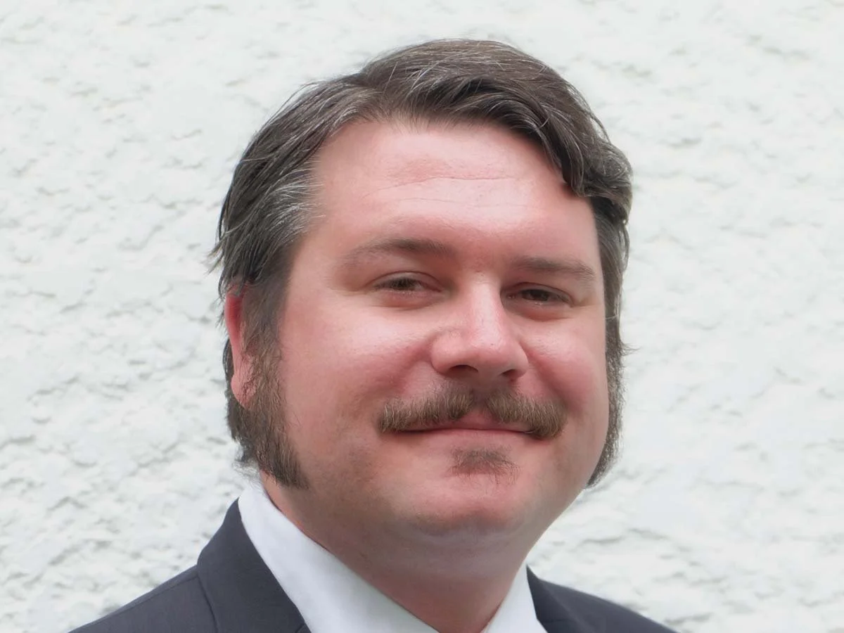 A man with dark hair, a mustache, and sideburns wearing a suit and tie, standing in front of a textured, off-white wall.