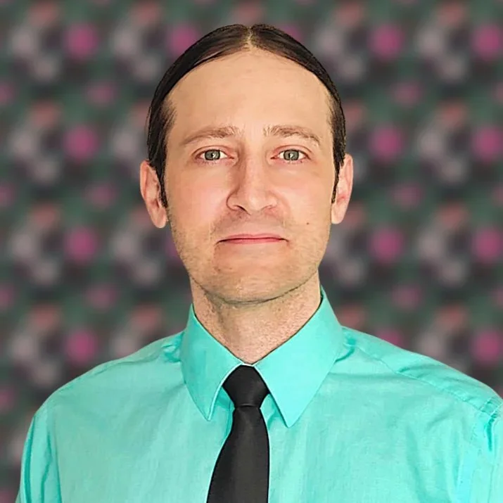 A man with dark hair, wearing a teal dress shirt and black tie, standing in front of a blurred background with pink and gray pattern.