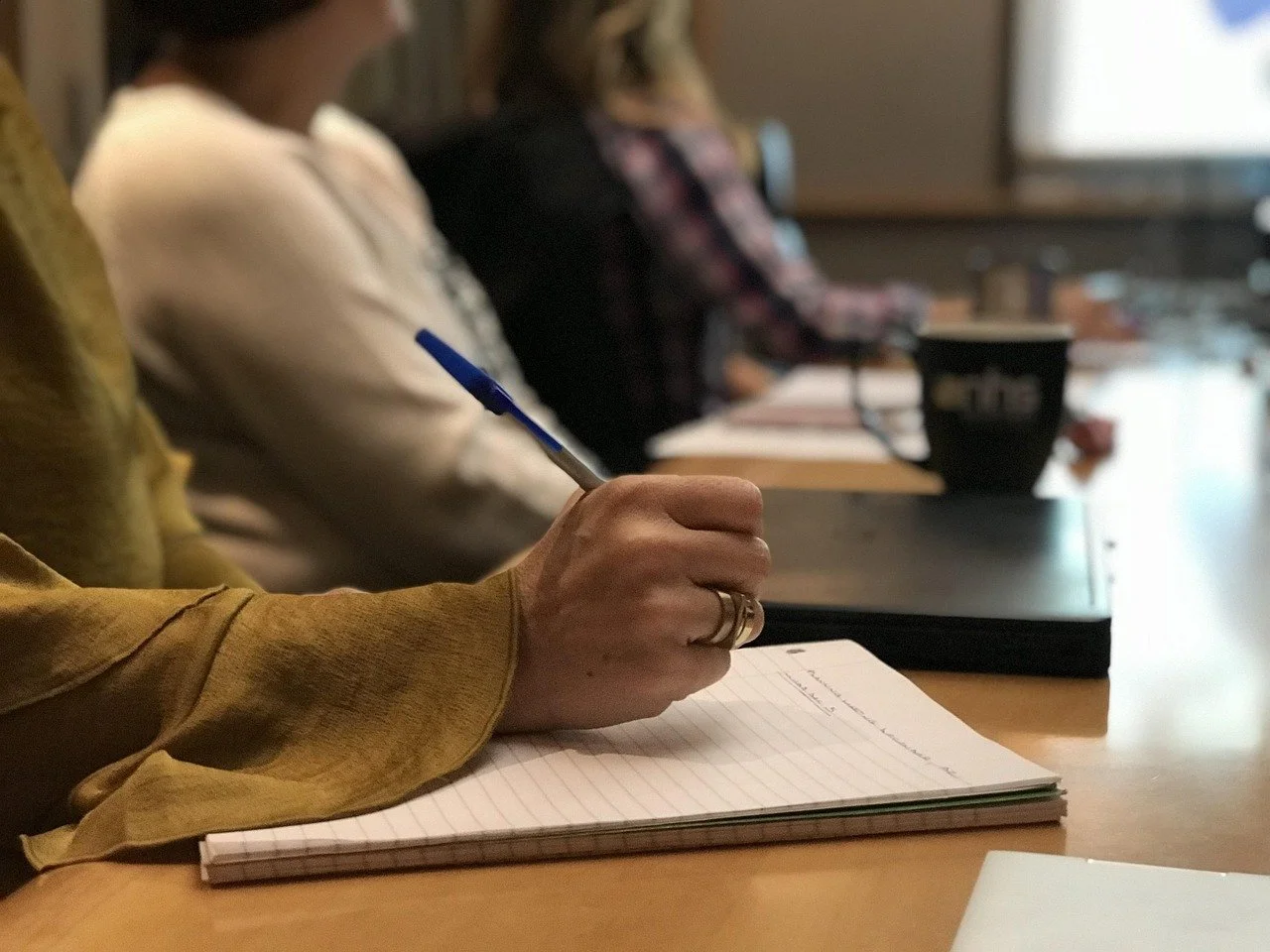 Close-up of a person's hand writing on a notepad with a blue pen, with other people sitting at a table in the background.