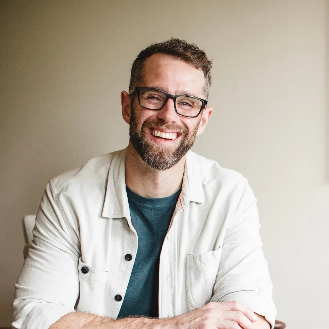 Smiling man with glasses and a beard, wearing a beige shirt, sitting at a table against a plain wall.