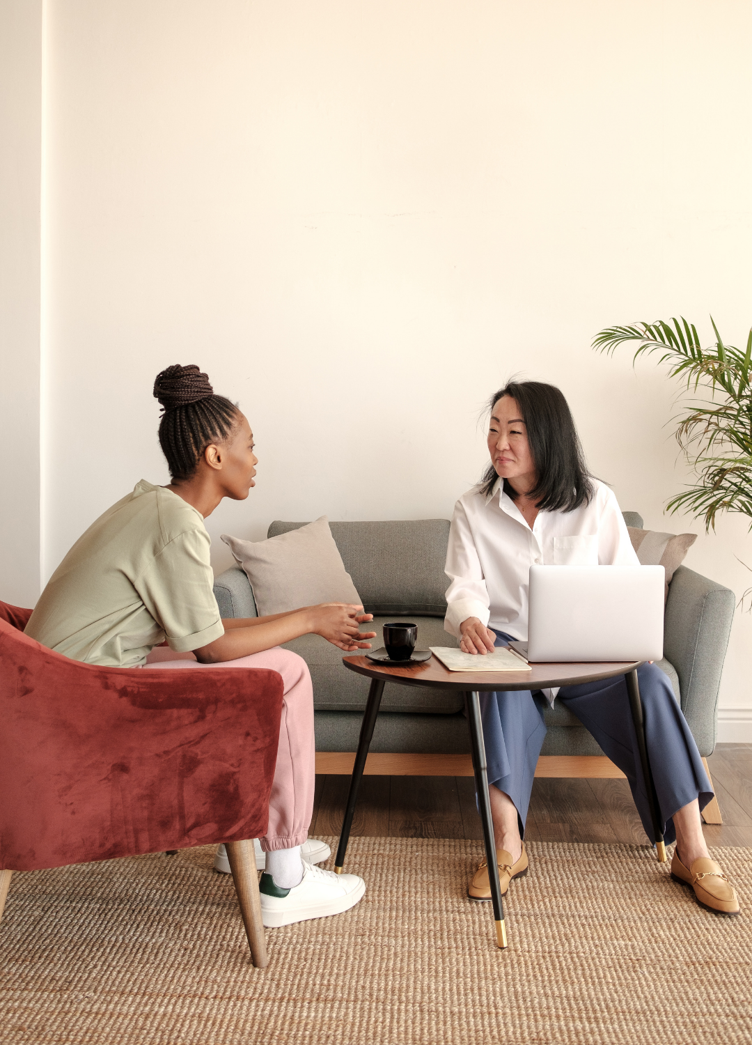 Two women sitting on opposite sides of a small round table, engaging in conversation. One woman has dark skin and braids, wearing a beige shirt and pink pants. The other woman has light skin, black hair, and is wearing a white shirt and blue pants. They have a laptop, notebook, and coffee cup on the table, with a sofa and a plant in the background.