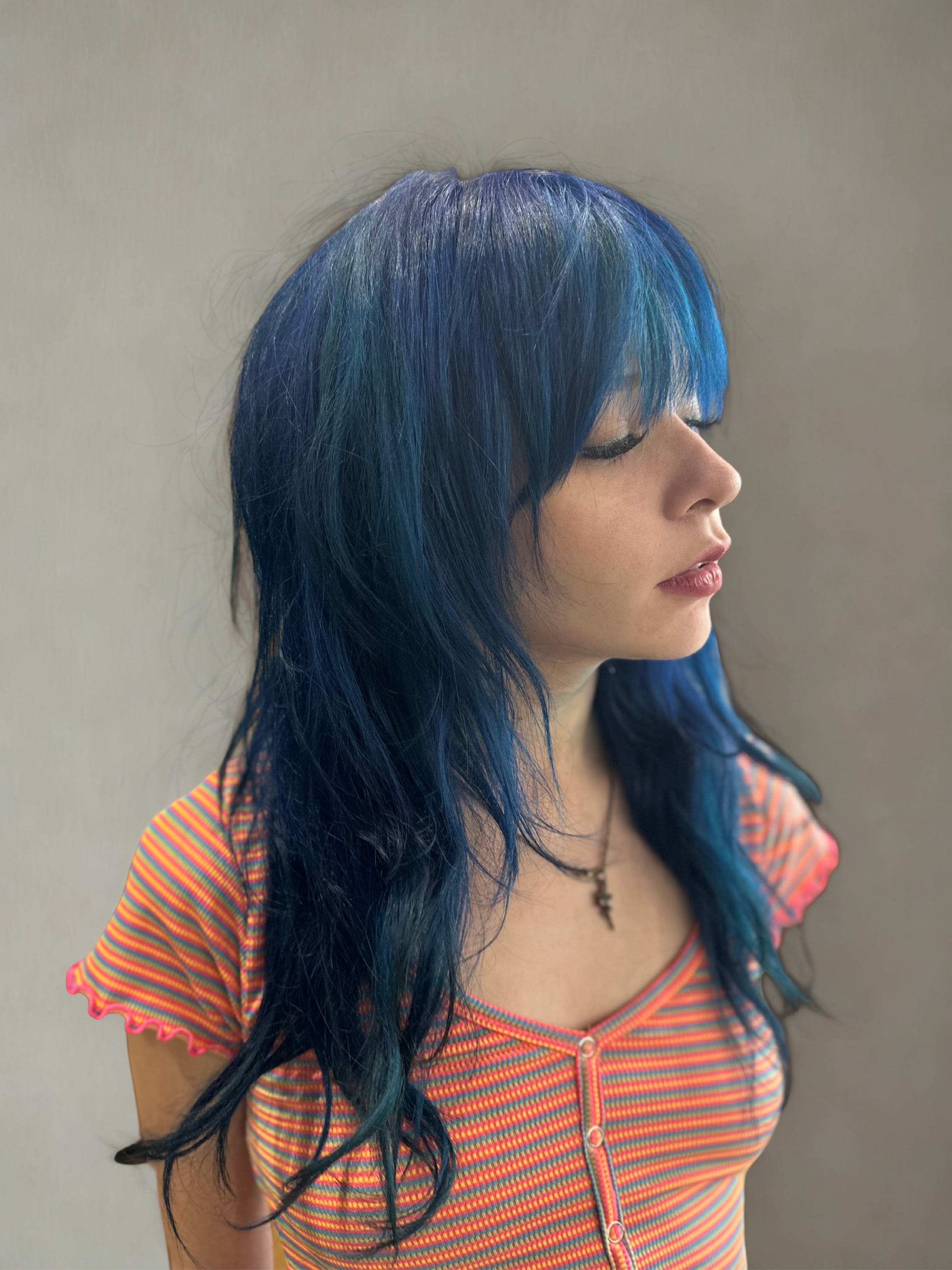 A woman with long, custom vivid wavy blue hair and bangs wearing a colorful striped top and a necklace, with her eyes closed and her head slightly tilted to the side against a plain background.