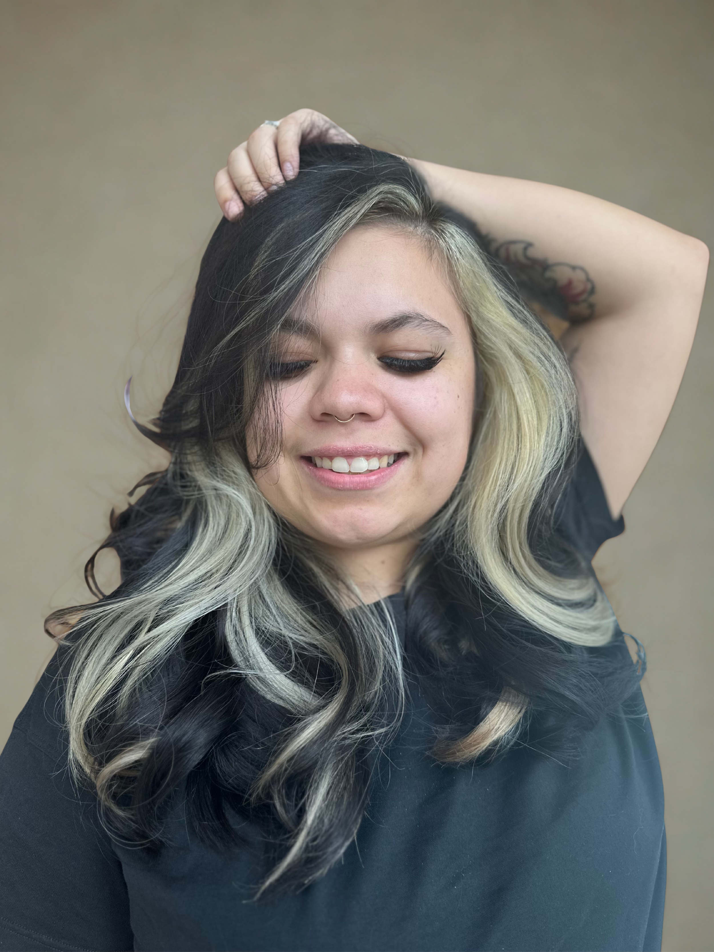 A woman with long wavy peekaboo black and blonde hair, smiling with eyes closed, wearing a black top and holding her hair back with one hand, against a plain beige background.
