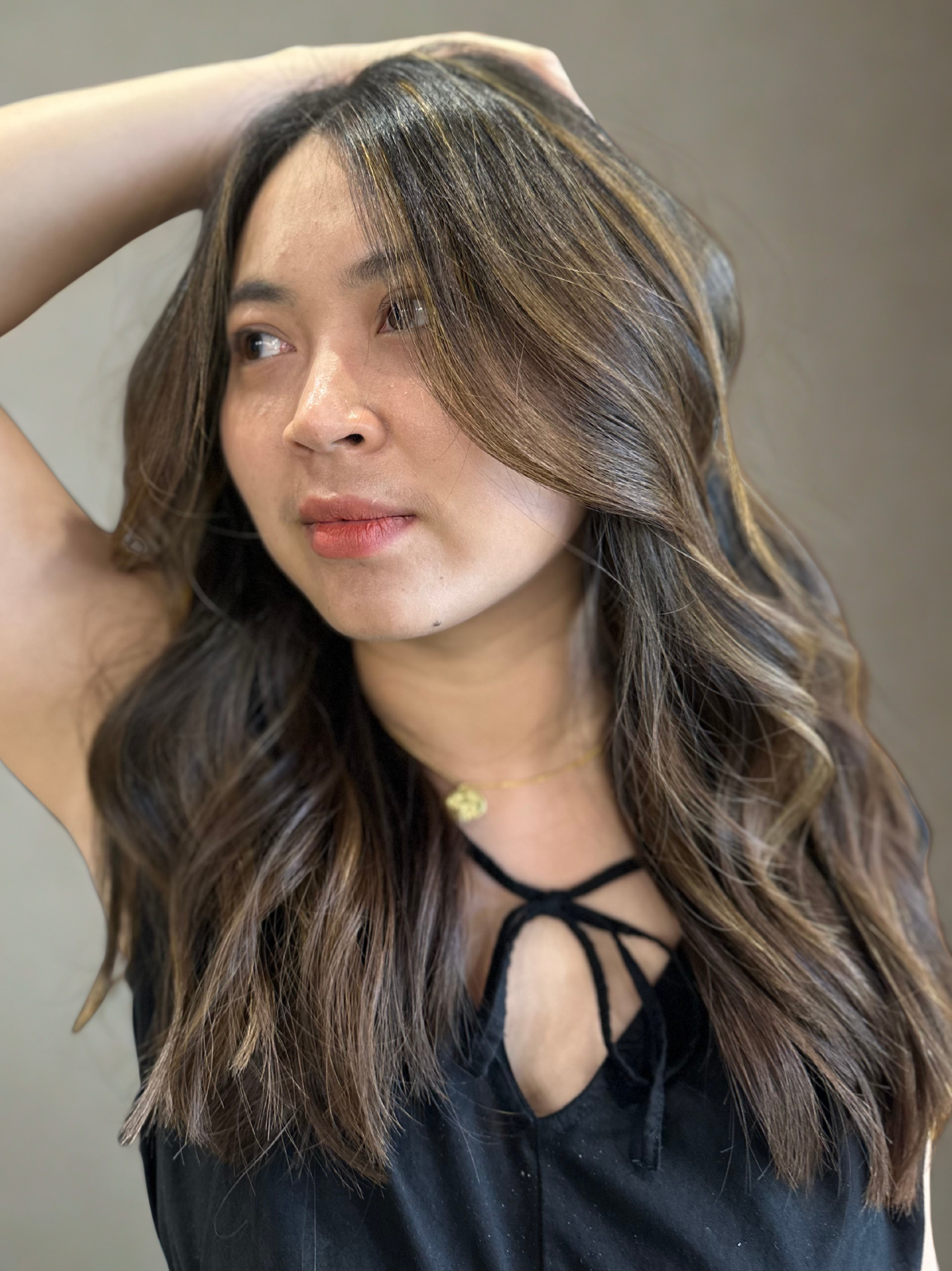 A woman with wavy brown hair and highlights, wearing a black top with a lace-up neckline and a gold necklace, looking to the side with her hand resting on her head.