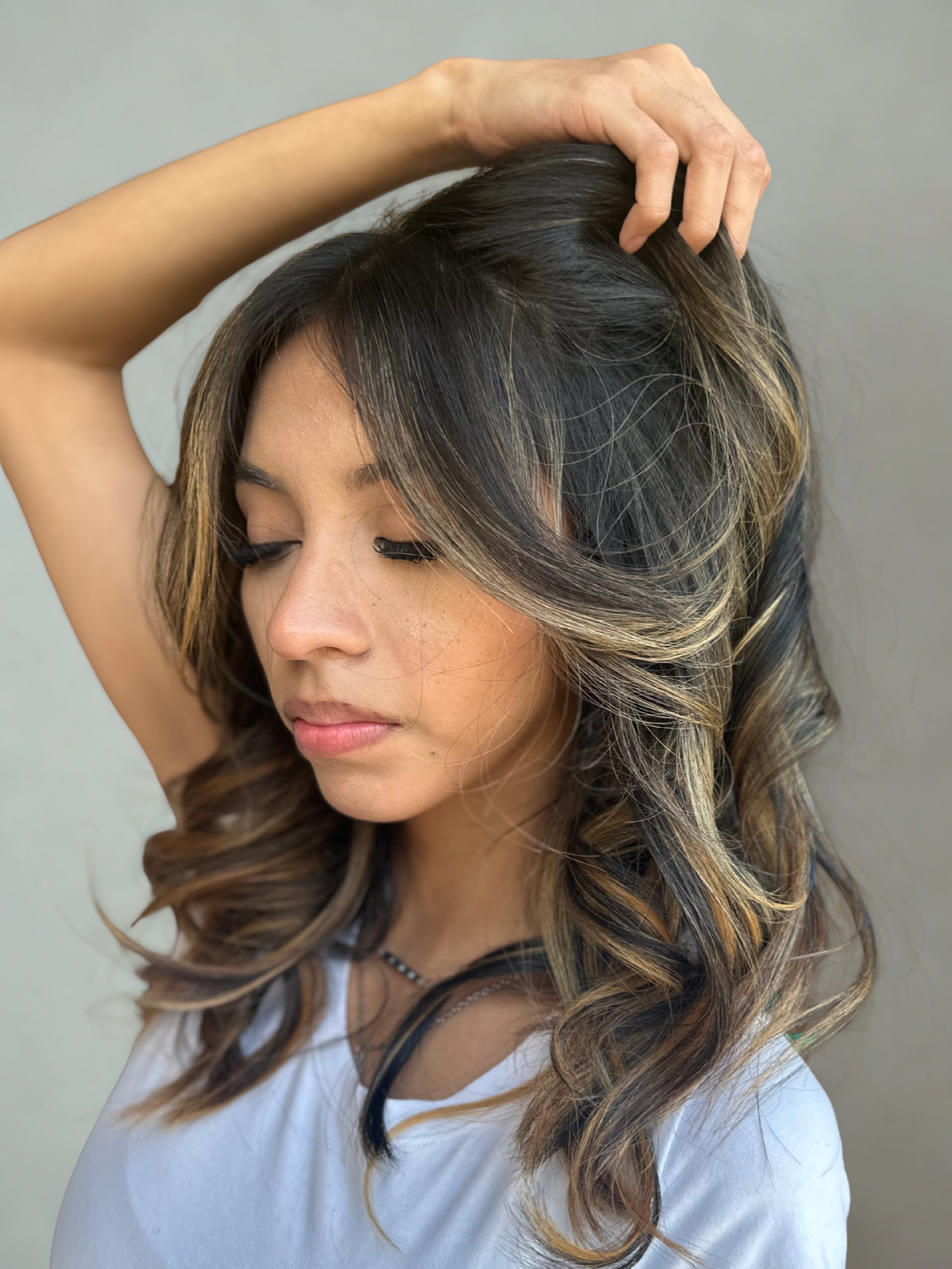Close-up of a woman with long, styled hair, brunette balayage color holding her head with one hand, eyes closed, wearing a white top and a necklace, against a neutral background.
