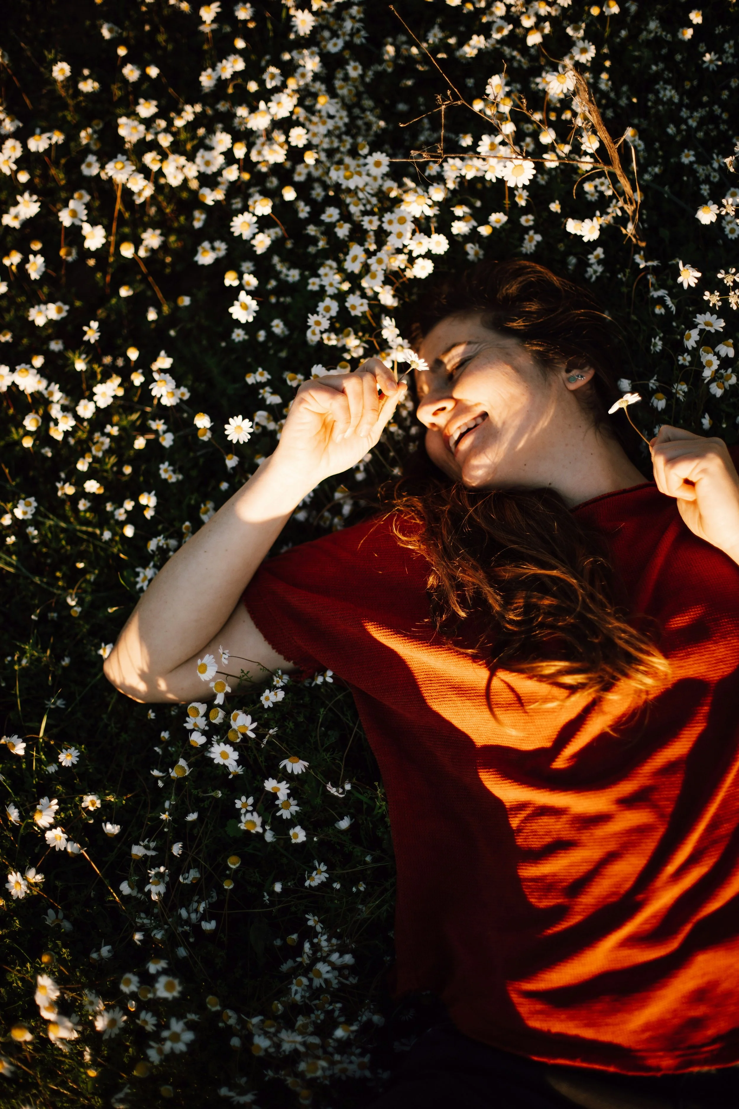 A woman with long dark hair lying on her back on a bed of small white daisies, smiling and holding daisies near her face in warm sunlight.