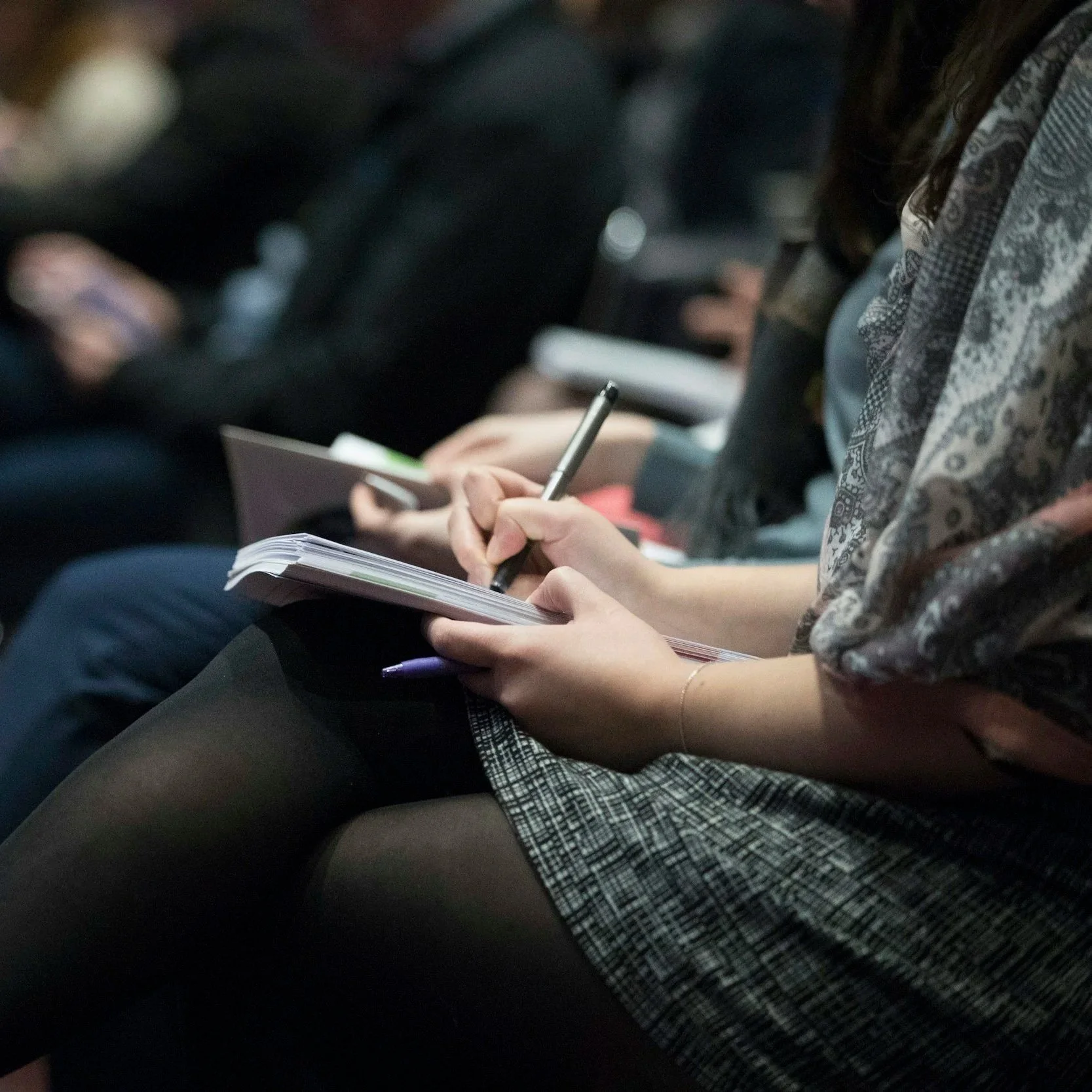 Close-up of a person's hands holding a notebook and a pen, sitting on a chair, with another person's arm visible, wearing a plaid skirt and a patterned scarf. The background includes other people in an indoor setting.