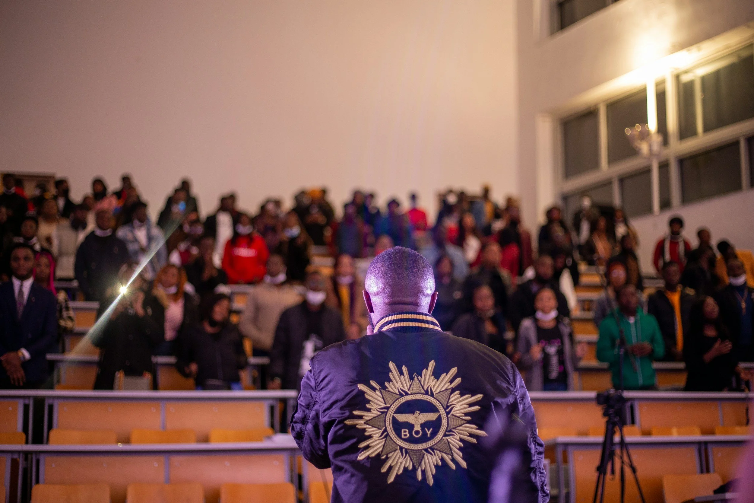 A person with a jacket displaying a large emblem reading 'BOY' on the back, facing an audience in an auditorium. The audience members are standing, and the auditorium is well-lit with large windows and a high ceiling.