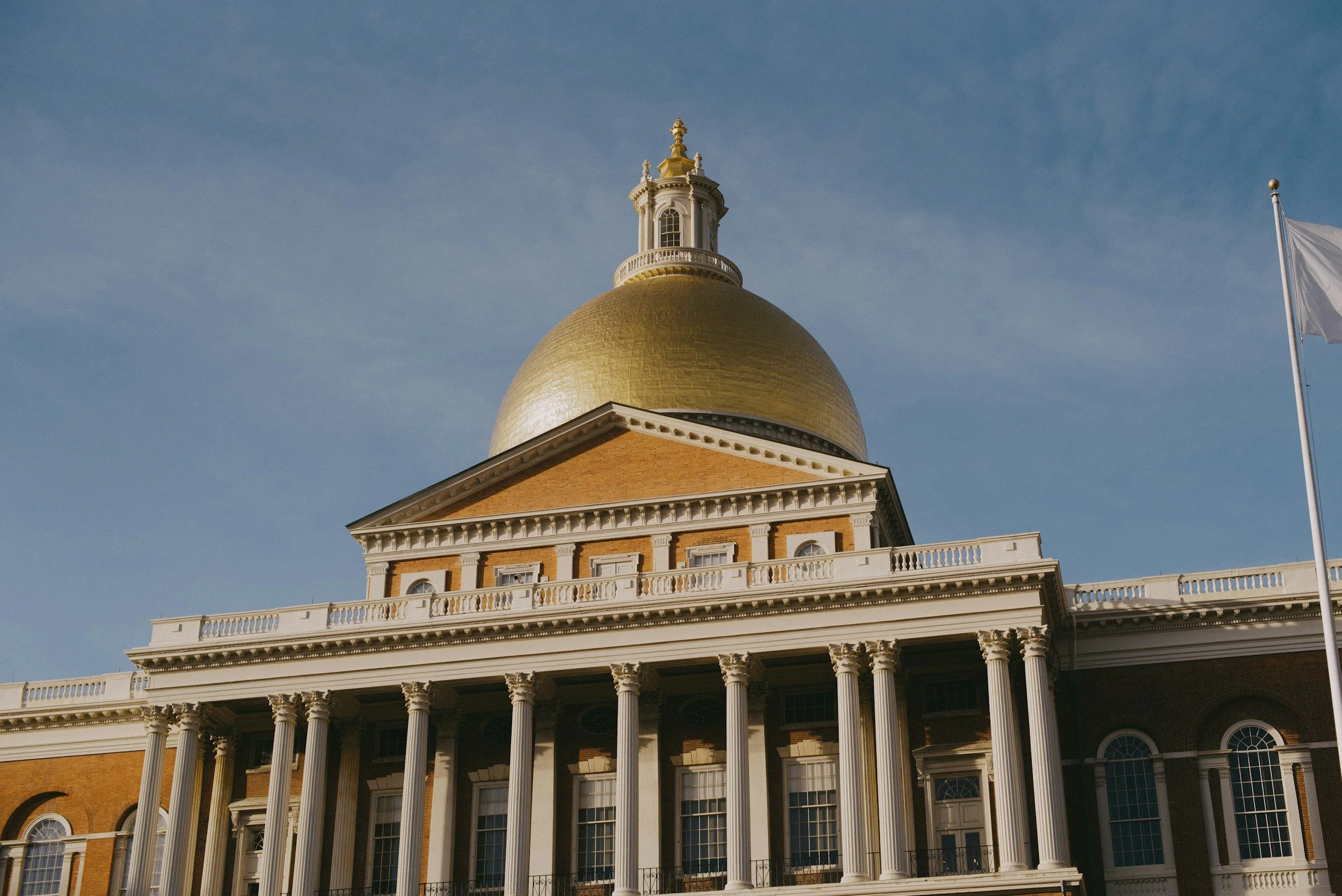 A historic building with a large golden dome, white columns, and a small tower at the top of the dome, under a clear blue sky, with a white flag on a pole to the right.