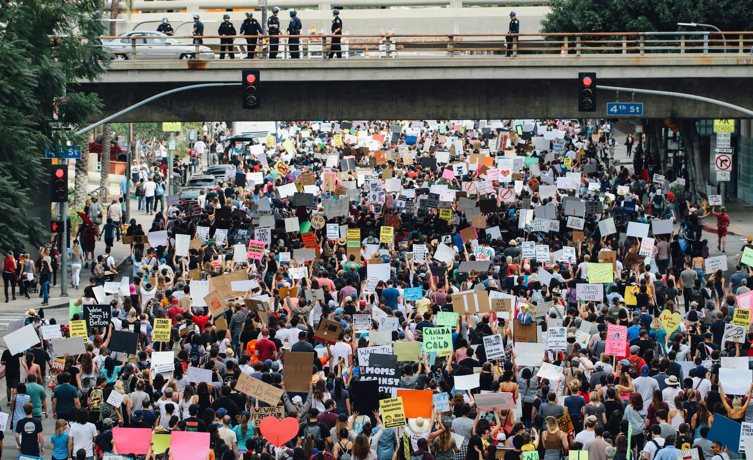 Large crowd of protesters holding signs on a city street, with police officers on a bridge above. The signs contain various messages for social and political causes.